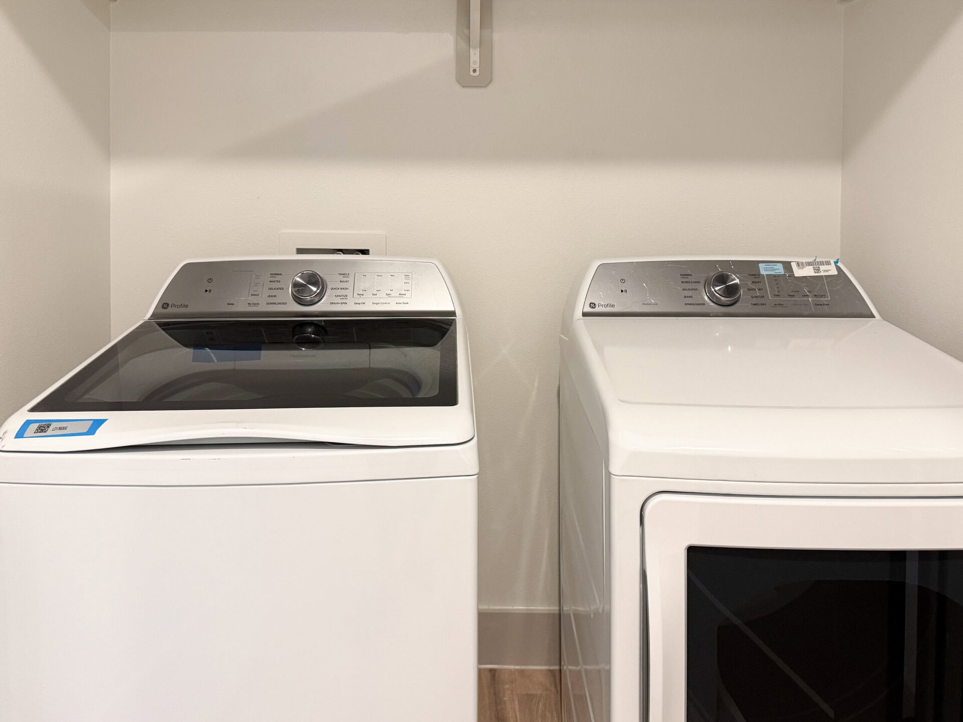 A washing machine and a dryer are positioned side by side in a laundry area. The washing machine on the left features a transparent lid and a control panel with various settings and a large dial. The dryer on the right has a similar control panel but lacks a transparent lid. Both appliances are white and have a modern design, with the washing machine displaying a label on its top. The background is a plain, light-colored wall.