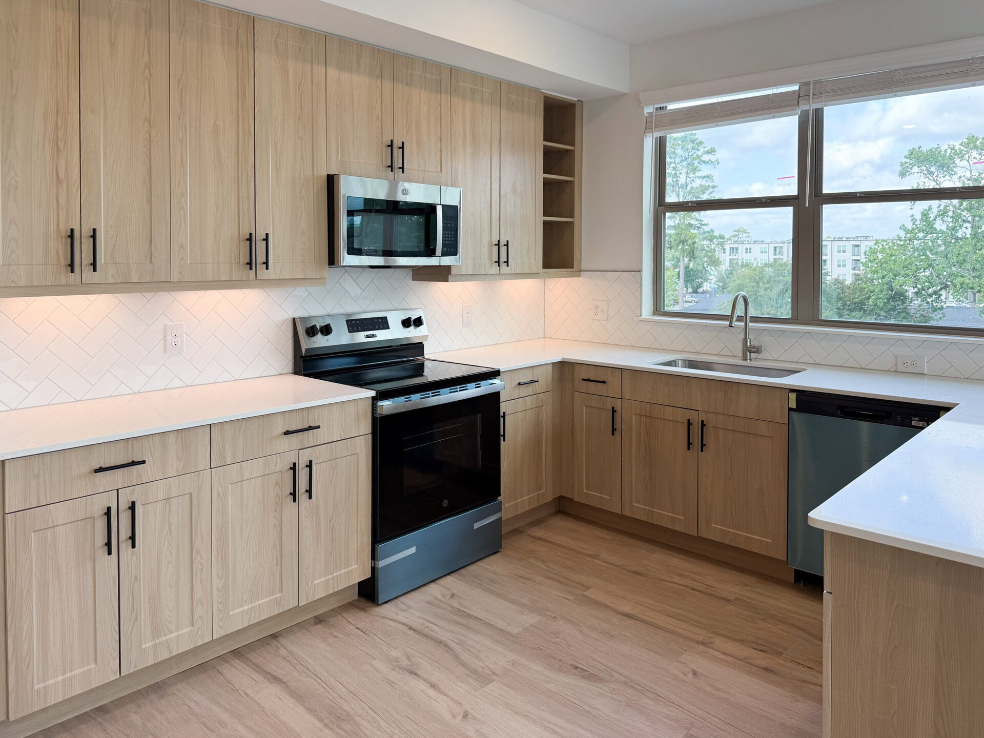 A modern kitchen featuring light wood cabinetry with black handles, a stainless steel microwave above a black electric stove, and a white countertop. The backsplash is designed with white herringbone tiles. A stainless steel sink is positioned under a window, providing natural light, while a dishwasher with a black front is located next to the sink. The flooring is light wood, enhancing the contemporary aesthetic.