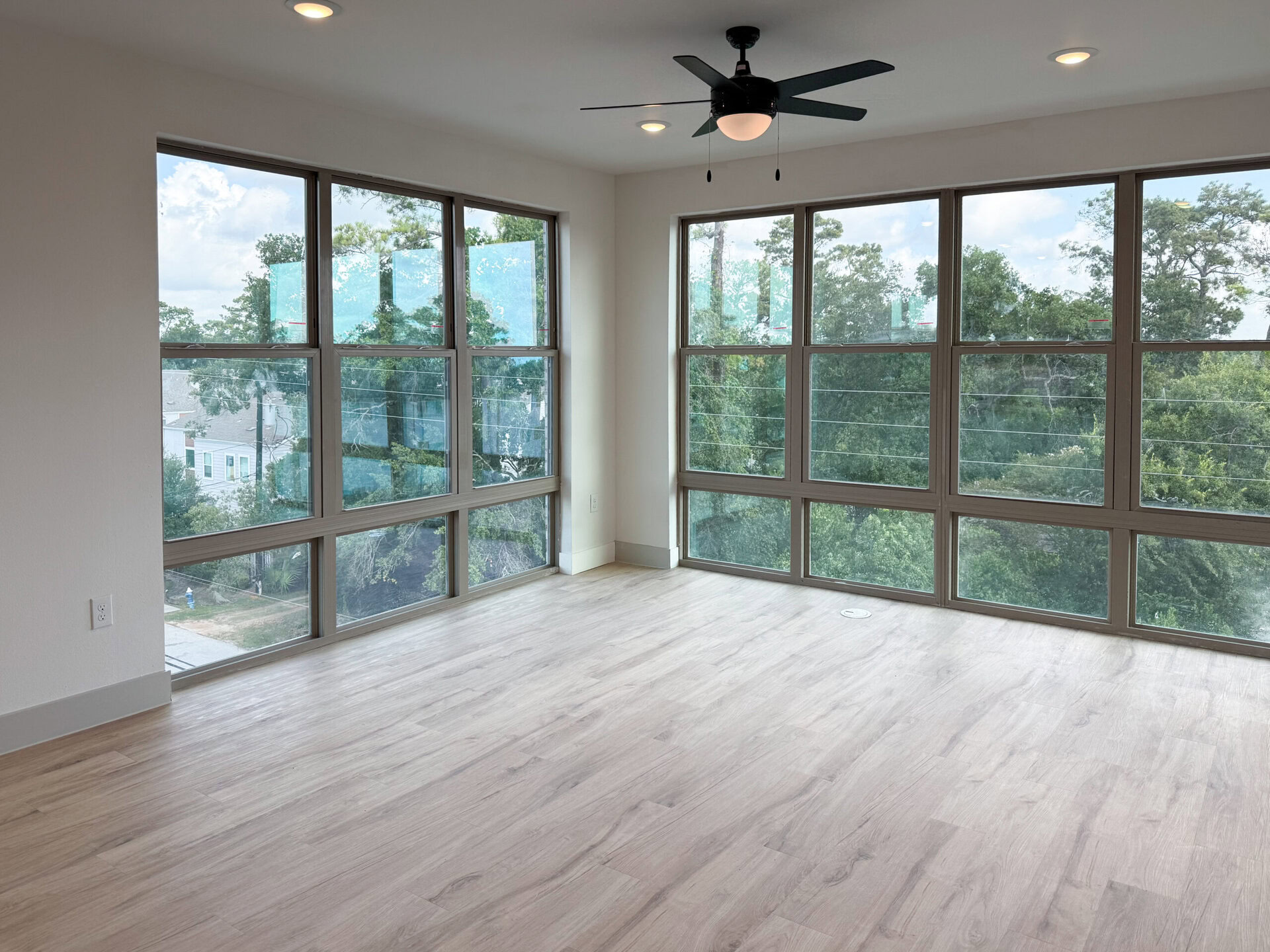 A spacious room features large windows that provide ample natural light and a view of greenery outside. The flooring is light-colored wood, and the walls are painted in a neutral tone. A ceiling fan is visible, adding to the modern aesthetic of the space. The overall atmosphere is bright and airy, ideal for a living area or workspace.
