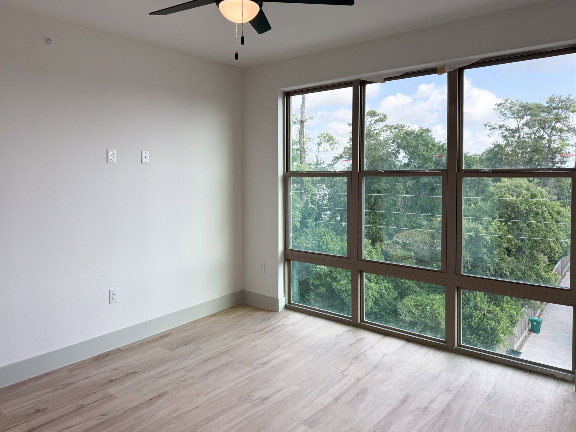 A bright, empty room features a large window with multiple panes, allowing natural light to flood in. The walls are painted a soft white, and the floor is covered with light-colored laminate. Two electrical outlets are visible on one wall, and outside the window, lush greenery and trees can be seen, along with a glimpse of a paved area and a green trash bin. The overall atmosphere is open and airy.