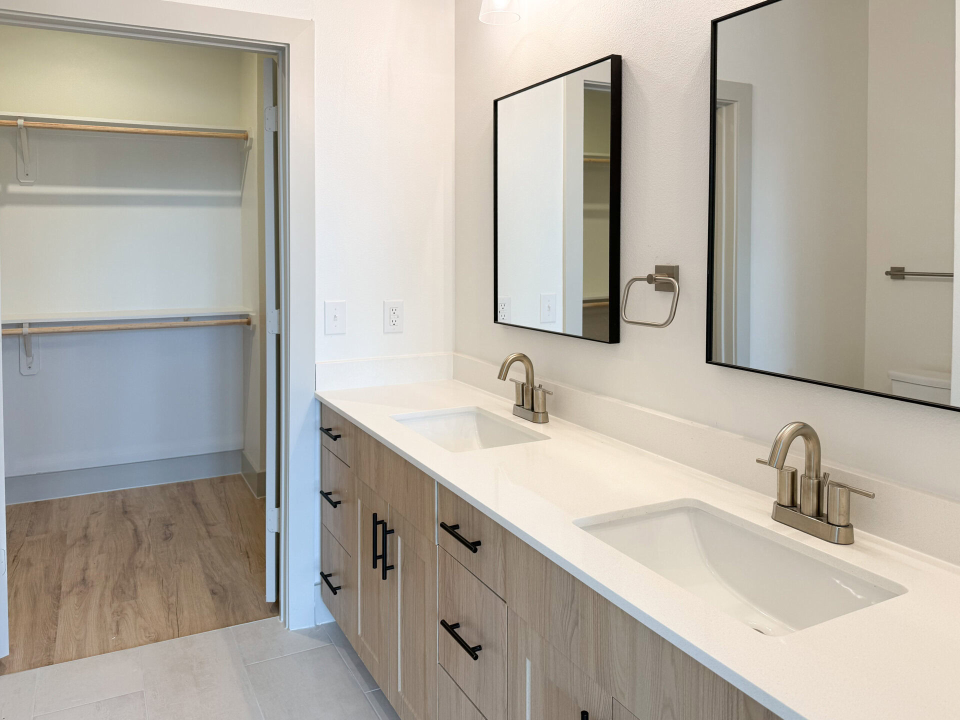 A modern bathroom features a double vanity with a white countertop and two rectangular sinks. Above each sink, there are rectangular mirrors framed in black. The cabinetry below is light wood with black handles. To the left, there is an open closet space with wooden shelves, and the flooring is a light wood laminate. The overall color scheme is neutral and contemporary.