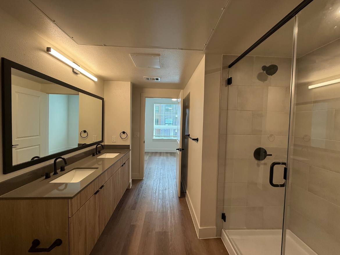 A modern bathroom features a double vanity with a sleek countertop and two sinks, accompanied by black faucets. Above the vanity, a large rectangular mirror is framed in black, with wall-mounted lighting. To the right, a glass shower enclosure showcases neutral-toned tiles. The floor is made of light wood, and a door leads to a well-lit room with windows, suggesting a spacious layout.