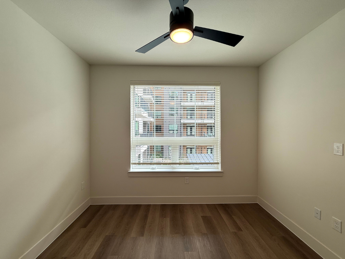 A small, empty room features light-colored walls and a wooden floor. A ceiling fan with a light fixture is mounted above. The room has a window with horizontal blinds, allowing natural light to enter, and offers a view of an adjacent building. There are electrical outlets on the walls, but no furniture or decorations are present.