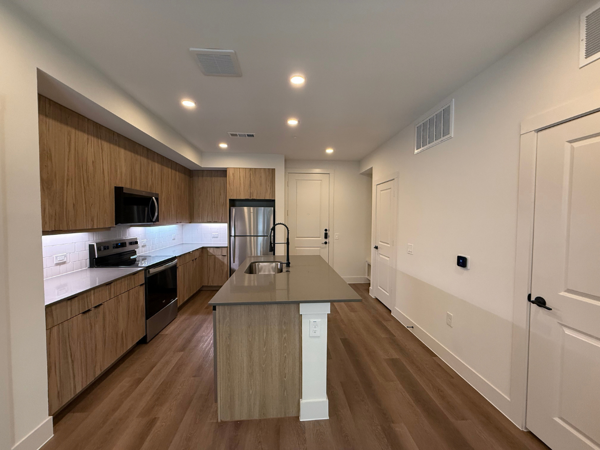A modern kitchen featuring light wood cabinetry and a sleek design. The kitchen includes stainless steel appliances, such as a refrigerator and an oven, with a black microwave above the stove. A large island with a gray countertop is positioned in the center, providing additional workspace. The backsplash is made of white subway tiles, and under-cabinet lighting adds a warm glow. The flooring is a light wood, and there are white walls with a door leading to another room. A thermostat is mounted on the wall.