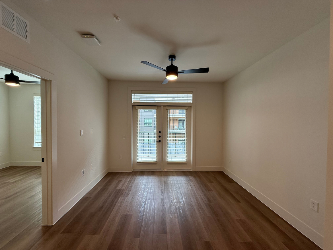 A spacious room with light-colored walls and a wooden floor. There is a ceiling fan with black blades and a light fixture. On one side, double doors lead to a small balcony, with blinds partially covering the windows. To the left, there is an open doorway leading to another room, which has a window with vertical blinds. The overall atmosphere is bright and modern.