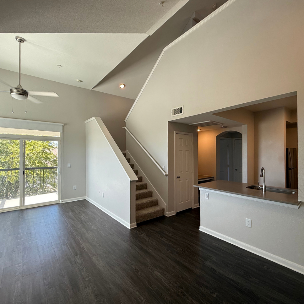 A spacious living area features a high ceiling with a ceiling fan, large windows allowing natural light, and a sliding glass door leading to a balcony. A staircase with carpeted steps ascends to an upper level. To the right, an open kitchen area includes a countertop with a sink, and there is a doorway leading to another room. The flooring is dark, resembling wood, while the walls are painted in a light neutral color.