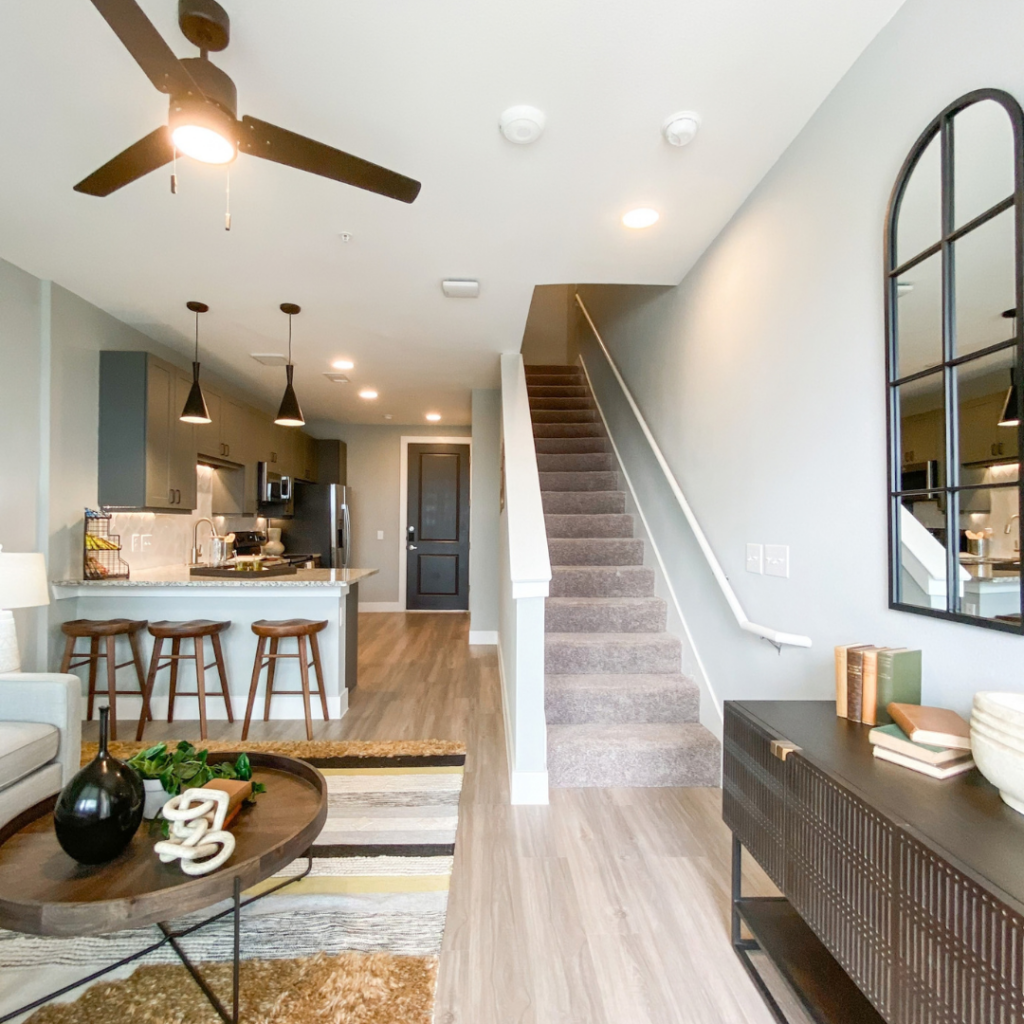 A modern living space features an open layout with a kitchen and living area. The kitchen, equipped with dark cabinetry and stainless steel appliances, includes a granite countertop with three wooden bar stools. A cozy seating area is adorned with a light-colored sofa and a round coffee table, which holds decorative items and a small plant. The floor is a mix of light wood and area rugs. A staircase with carpet leads to an upper level, while a large mirror with an arched frame hangs on the wall. Soft lighting from ceiling fixtures enhances the inviting atmosphere.