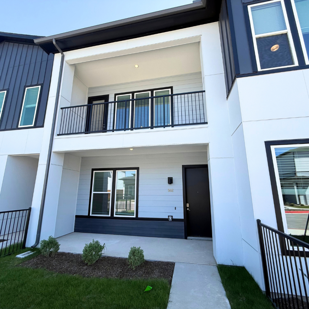 A modern two-story building features a clean, contemporary design with a mix of white and dark siding. The ground floor has a small porch area with a concrete walkway leading to a black front door. Above, a balcony with a black metal railing overlooks the entrance. Large windows on both levels allow natural light to enter, and small shrubs are planted in the landscaped area in front of the building. The overall aesthetic is sleek and inviting.