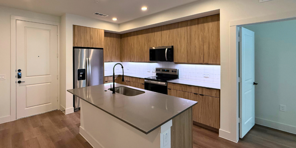 A modern kitchen featuring light wood cabinetry and stainless steel appliances. The kitchen island has a gray countertop with a sink and a stylish black faucet. A refrigerator and a microwave are integrated into the cabinetry, while an electric stove is positioned beneath a vent hood. The backsplash consists of white subway tiles, and the space is well-lit with recessed lighting. A door leads to another room, and the entrance to the kitchen is visible on the left. The flooring is a warm wood tone, enhancing the contemporary feel. Learn all about a Smart City Exclusive, and see how you could save money on rent!