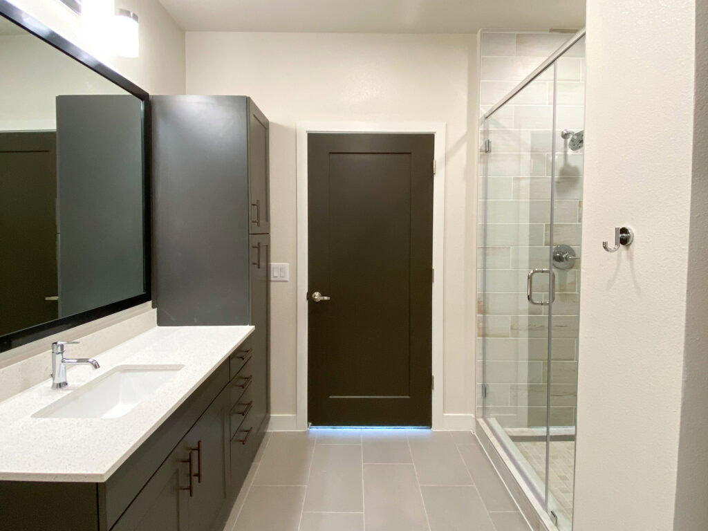 A modern bathroom features a sleek, dark cabinet with multiple drawers and a white countertop. A rectangular sink is integrated into the countertop, accompanied by a polished chrome faucet. To the right, a glass shower enclosure showcases light-colored tiled walls. A dark door leads out of the bathroom, and a small chrome hook is mounted on the wall nearby. The overall color scheme combines dark cabinetry with light walls and flooring, creating a contemporary aesthetic.