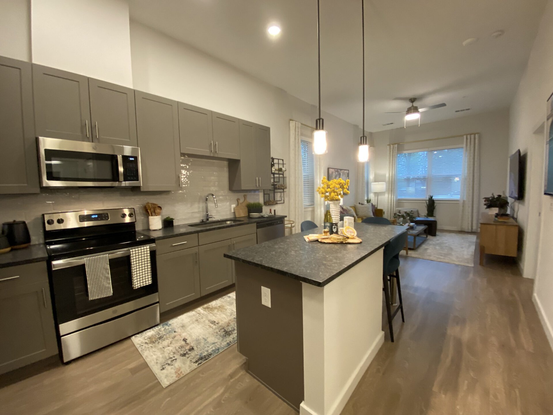 A modern kitchen features gray cabinetry and stainless steel appliances, including a microwave and an oven. The countertop is dark and extends into a bar area with high stools. A decorative arrangement of yellow flowers sits on the counter alongside snacks. In the background, a cozy living area is visible, with a sofa, coffee table, and large windows adorned with light curtains, allowing natural light to filter in. The flooring is a light wood, contributing to the contemporary aesthetic.