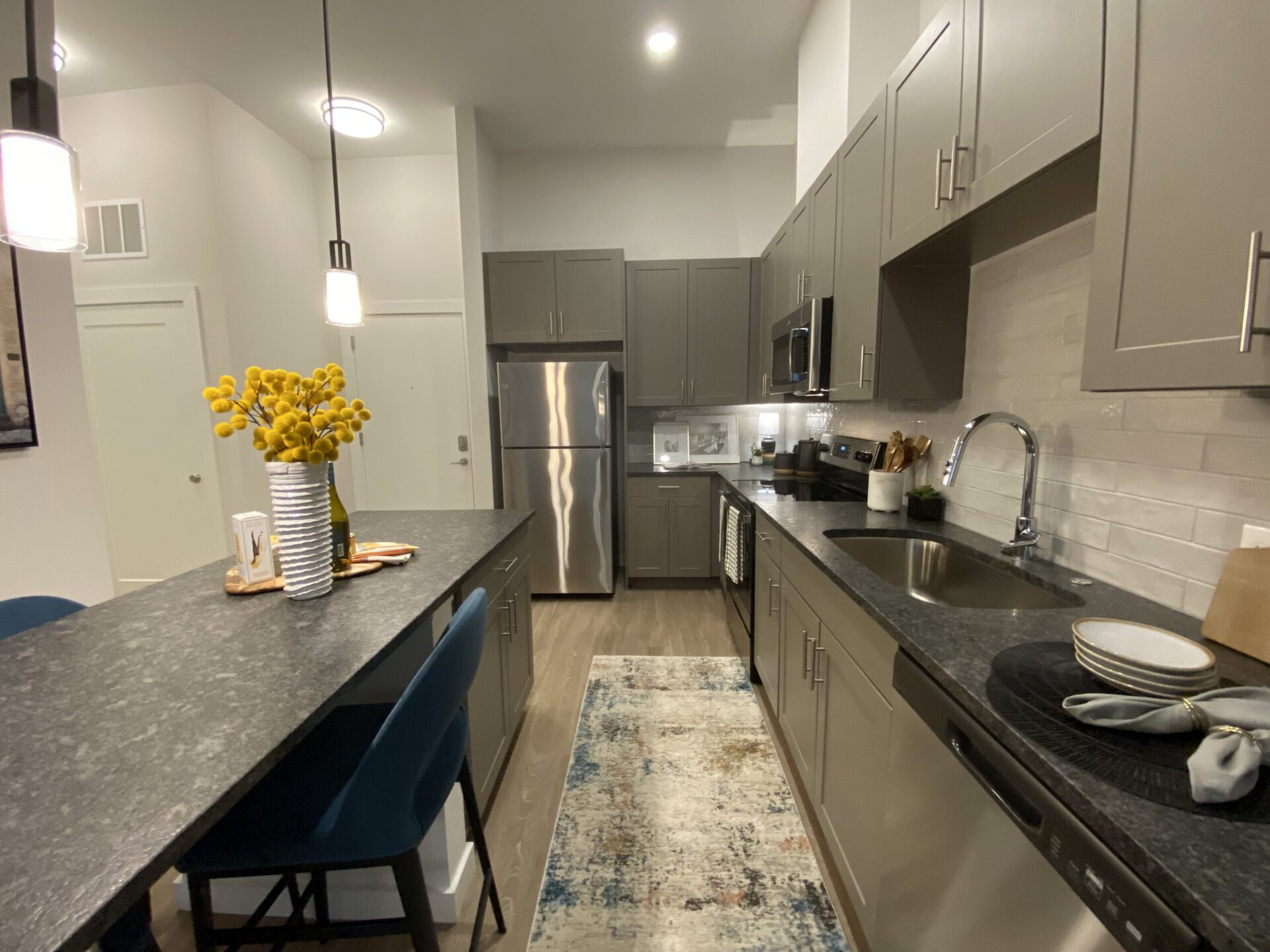 A modern kitchen featuring gray cabinetry and a dark countertop. The stainless steel refrigerator is positioned against the left wall, with an oven and microwave above it. A sink with a sleek faucet is located on the right side, next to neatly arranged plates and a folded napkin. A decorative vase filled with yellow flowers sits on the countertop, accompanied by a cutting board and some snacks. The floor is covered with a colorful area rug, and the space is illuminated by pendant lights.
