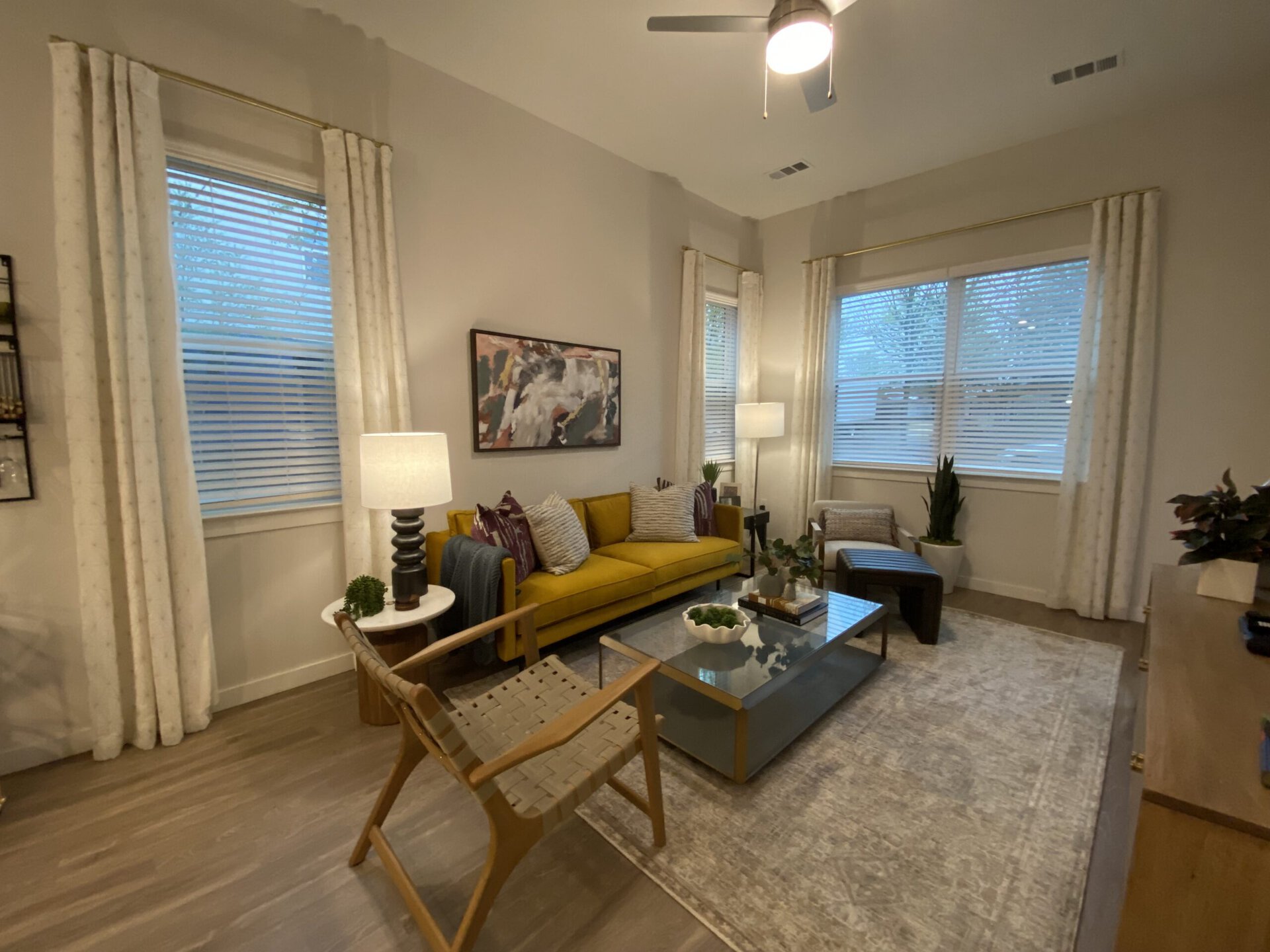 A cozy living room features a bright yellow sofa adorned with patterned throw pillows. A glass coffee table sits in front, displaying decorative items and a small plant. To the side, a wooden chair with a woven seat complements the space. Two large windows with white blinds allow soft light to filter in, and light-colored curtains frame them. A floor lamp and a small potted plant add warmth to the room, which has light wood flooring and a neutral rug. A piece of abstract art hangs on the wall, enhancing the modern aesthetic.