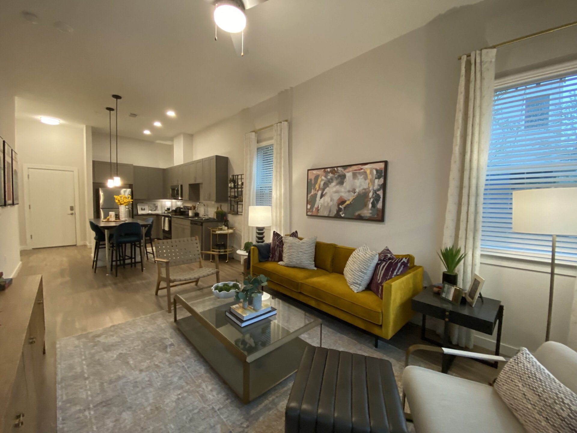 A modern living space features a vibrant yellow sofa adorned with patterned cushions, positioned in front of a glass coffee table. To the left, a stylish armchair complements the decor. The kitchen area, visible in the background, has sleek gray cabinetry and stainless steel appliances. A dining table with blue chairs sits nearby, topped with a vase of yellow flowers. Natural light filters through sheer curtains, illuminating the room, which also includes a floor lamp and decorative artwork on the walls.
