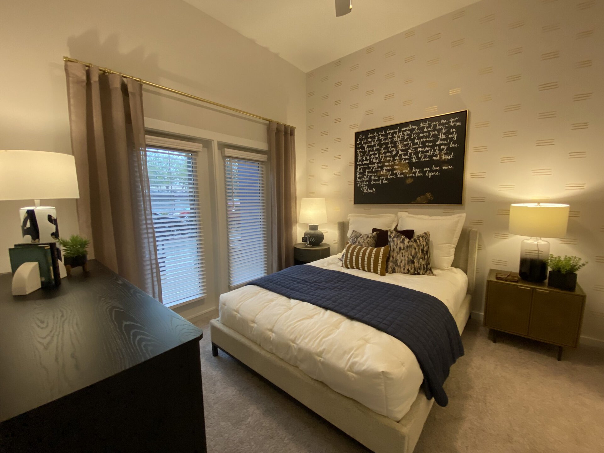 A cozy bedroom features a neatly made bed with white bedding and a navy blue quilt. Decorative pillows in various patterns and colors are arranged on the bed. To the left, a dark wooden dresser holds a small plant and a lamp with a unique design. Large windows with light brown curtains allow natural light to filter in, while a bedside table on the right displays a clock and a small plant. The wall behind the bed is adorned with a large black artwork featuring white handwritten text, adding a personal touch to the space.