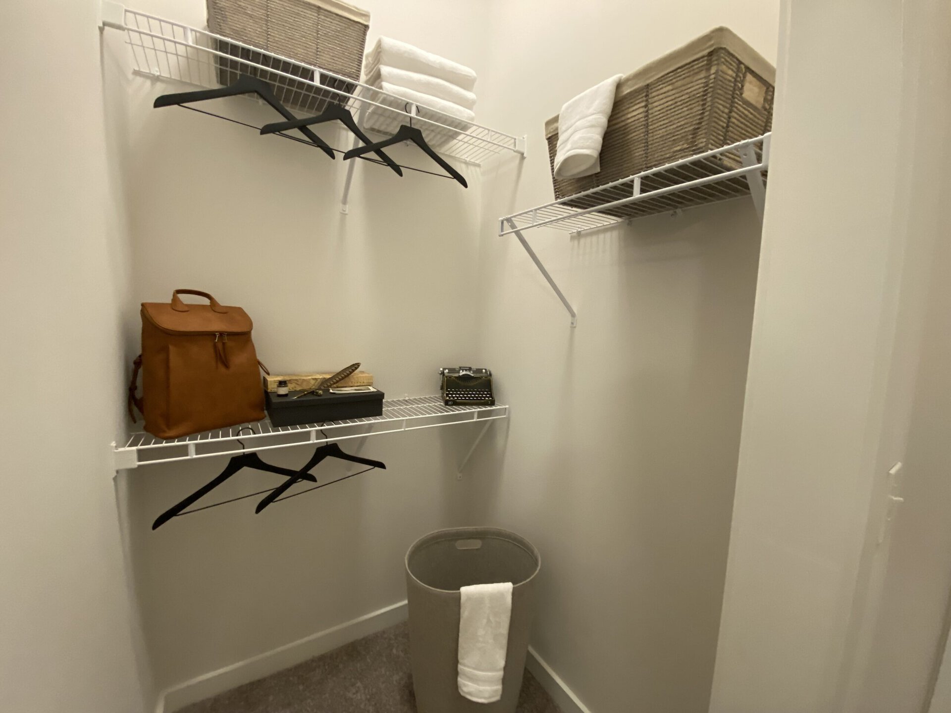 A small closet features white wire shelving with several black hangers. On the left side, a brown leather backpack rests on the floor next to a gray laundry basket. Above, a black box with a feather pen and a vintage typewriter are displayed on the shelf. Rolled white towels are neatly stacked in a woven basket on the top shelf. The walls are painted a light color, creating a bright and organized space.