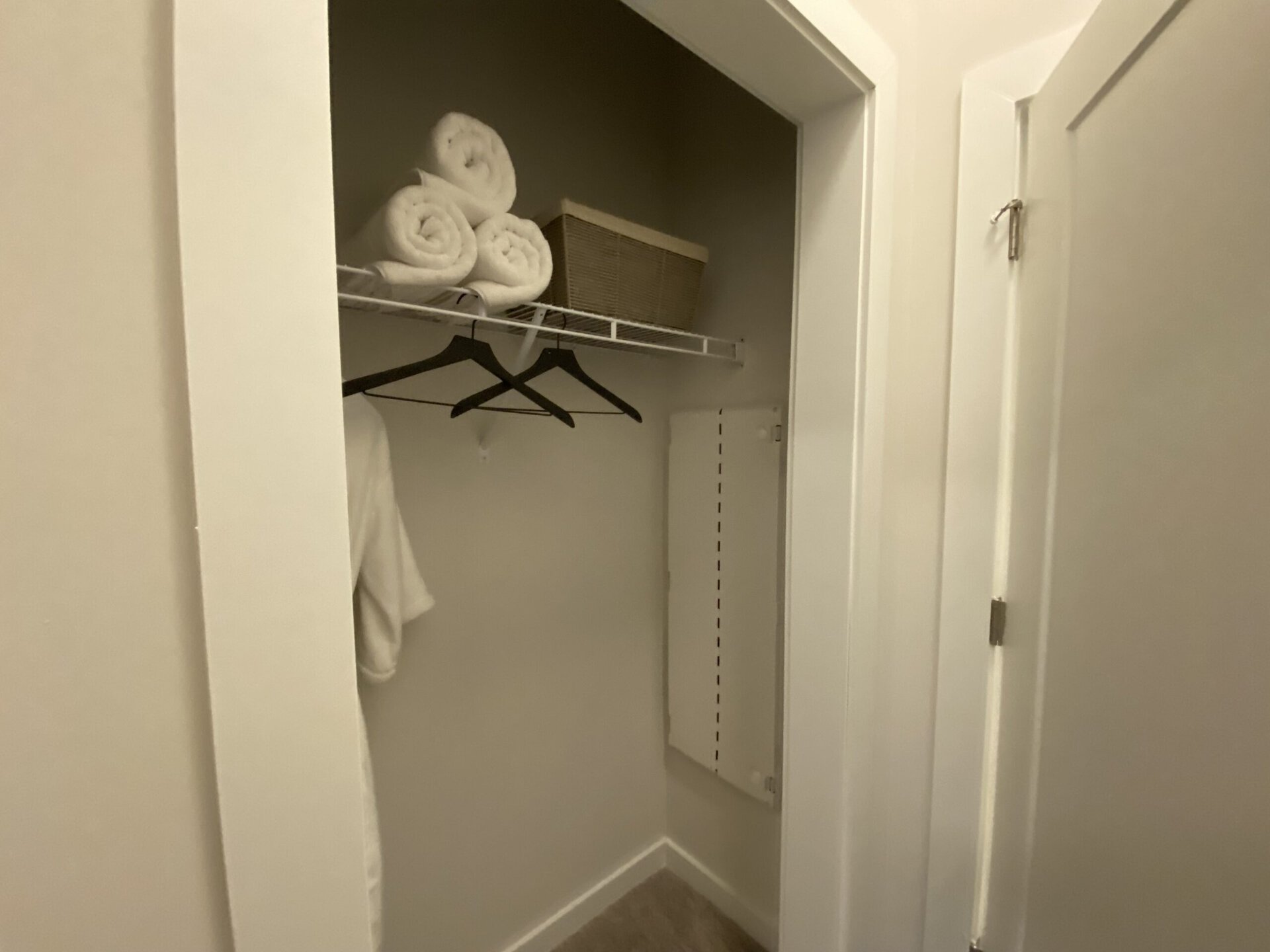 A small closet features a shelf with neatly rolled white towels stacked on top. Below, two black hangers are visible, one holding a white robe. To the side, there is a woven basket and a white panel with vertical lines, likely a folding door or storage unit. The walls are painted in a light neutral color, creating a clean and organized appearance.