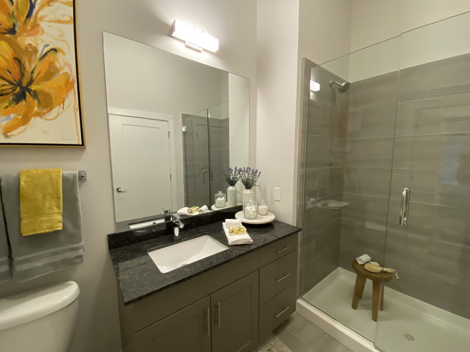 A modern bathroom features a sleek design with a large mirror above a dark granite countertop. The countertop holds neatly arranged toiletries, including a few glass containers and a small white towel. A pair of towels, one yellow and one gray, hang on a rack. The shower area is enclosed with clear glass, showcasing gray tiled walls and a wooden stool inside. A vibrant yellow flower painting adds a pop of color to the neutral-toned walls.