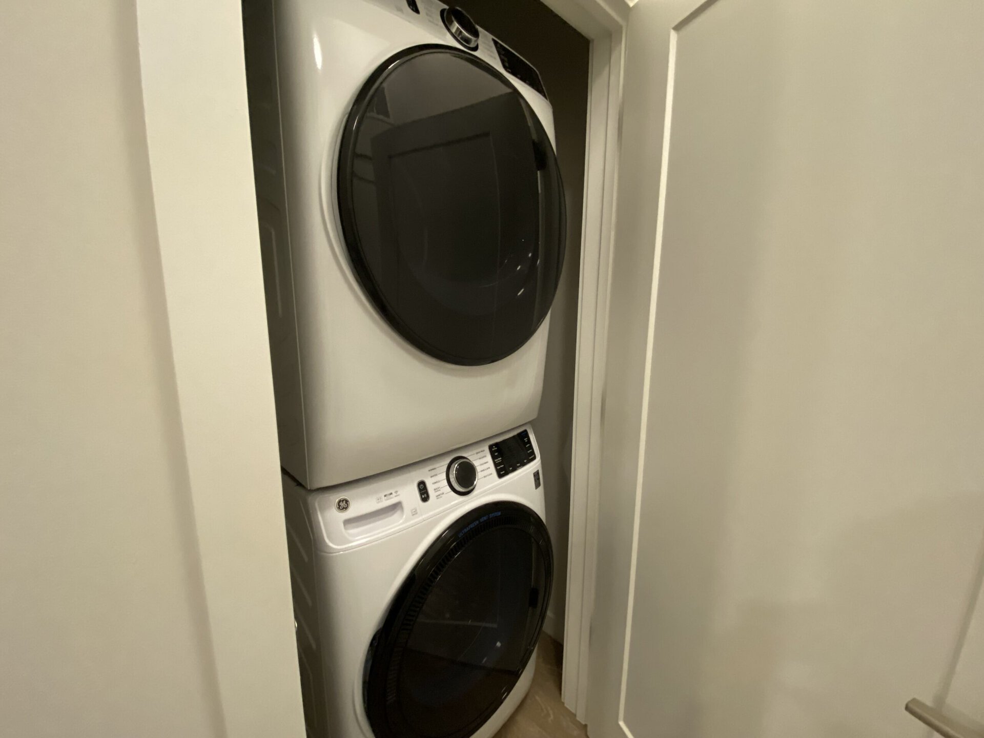 A stacked washer and dryer unit is positioned in a narrow space. The washer is located at the bottom, featuring a control panel with various settings and a circular door. The dryer, situated above the washer, has a large circular door as well. The appliances are white and have a modern design, with a light-colored wall on one side and a door frame visible on the other. The overall setting appears to be a compact laundry area.