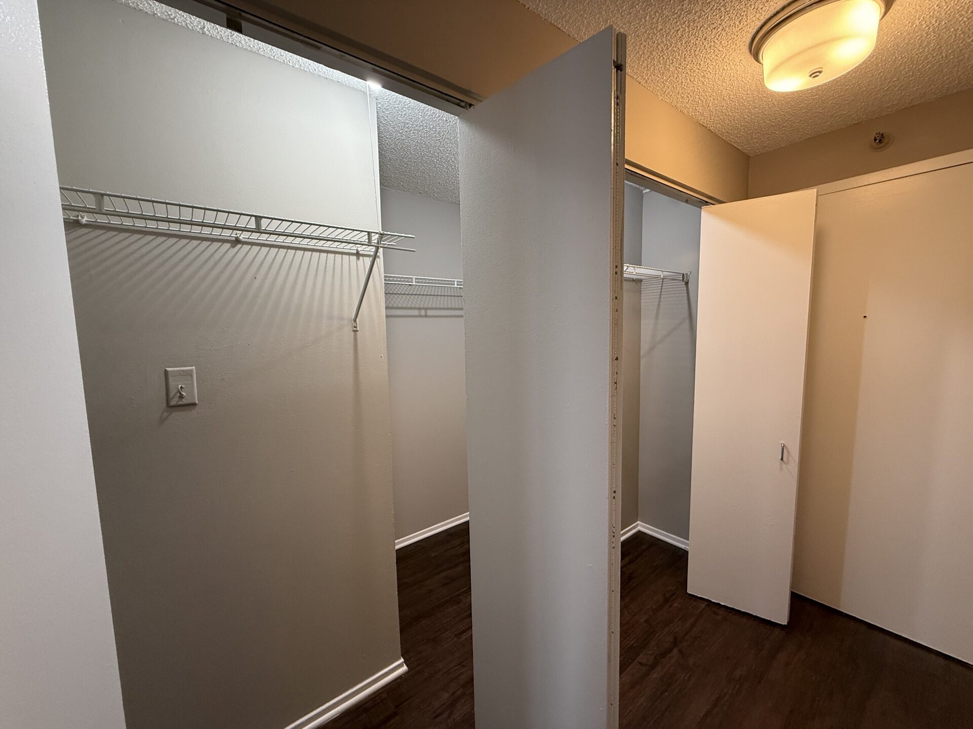 A spacious closet area is shown, featuring two separate sections. Each section has a wire shelving unit mounted on the wall, providing storage space. The walls are painted in a light gray color, and the flooring appears to be a dark wood laminate. One section has an open door, while the other is partially visible, creating a sense of depth in the space. The overall lighting is soft, enhancing the neutral tones of the room.