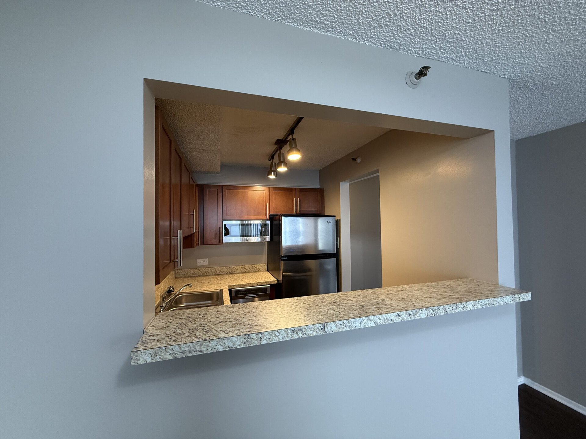 A modern kitchen is visible through an open wall cutout. The kitchen features wooden cabinetry, stainless steel appliances including a refrigerator and microwave, and a granite countertop. Above the countertop, there are three pendant lights providing illumination. The wall behind the kitchen is painted in a soft beige color, contrasting with the light gray walls surrounding the opening.