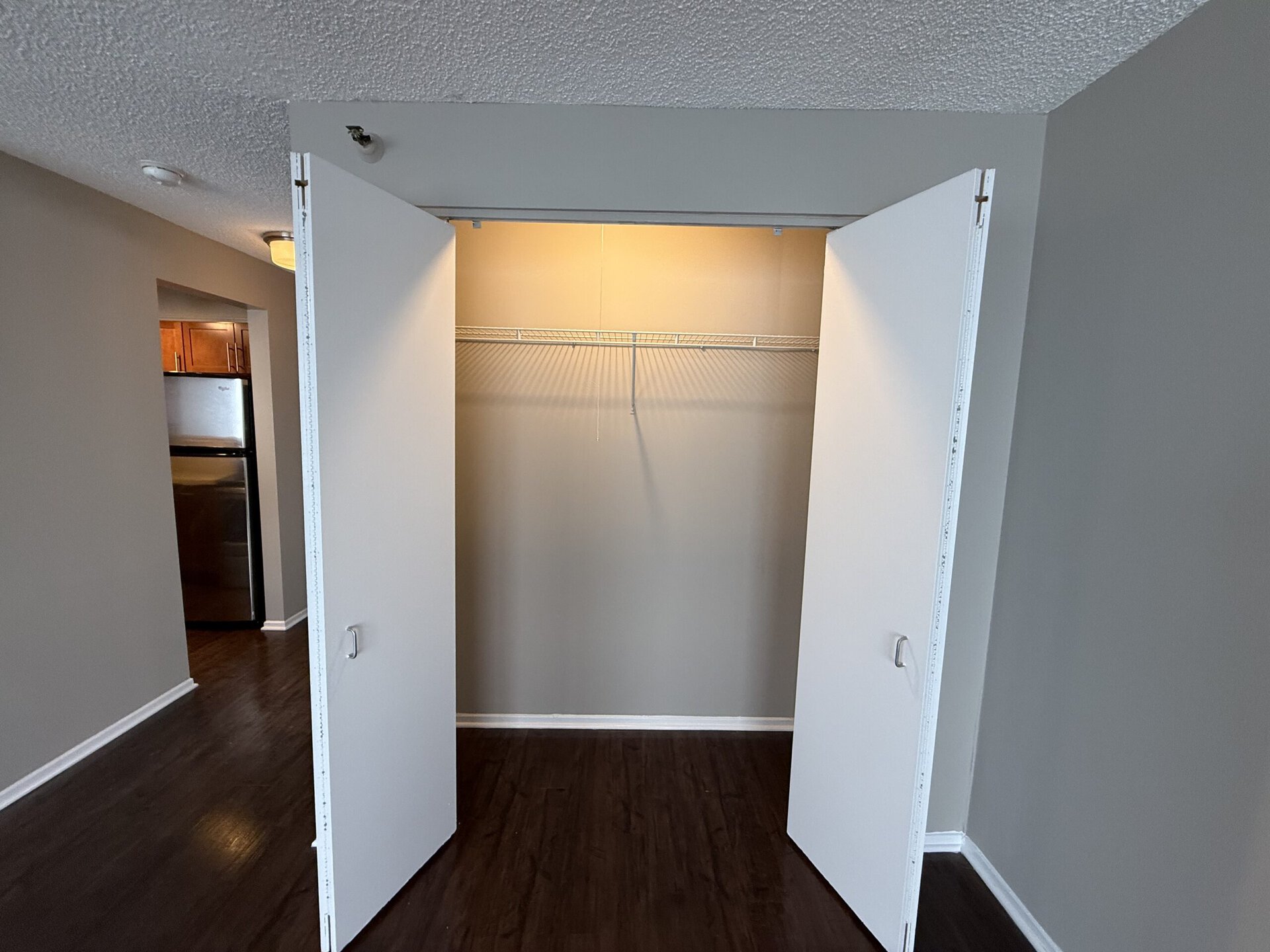 A spacious closet with double white doors is shown, opened to reveal an empty interior. The back wall is painted a light color, and a wire shelf runs along the top, providing storage options. The floor is dark wood, and there is a glimpse of a kitchen area with wooden cabinets and a black refrigerator visible in the background. The overall lighting is bright, emphasizing the clean and uncluttered space.