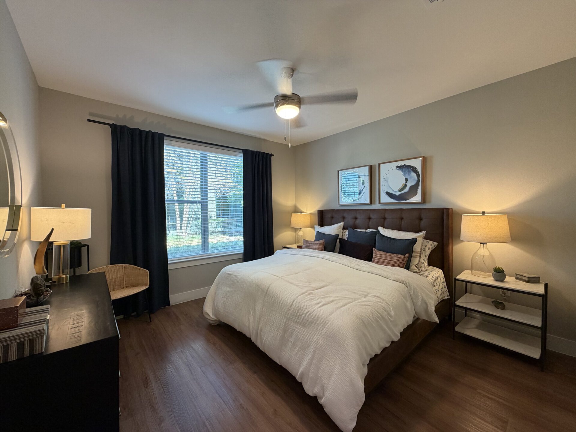A cozy bedroom features a large bed with a tufted brown headboard, dressed in a white comforter and an array of decorative pillows in various colors. To the left, a window with blinds is flanked by dark curtains, allowing natural light to filter in. A bedside table with a light lamp and a small plant sits next to the bed. Across from the bed, a black dresser holds decorative items, and a woven chair adds a touch of texture to the space. Two framed artworks hang above the bed, enhancing the room's aesthetic. The flooring is a warm wood tone, contributing to the inviting atmosphere.