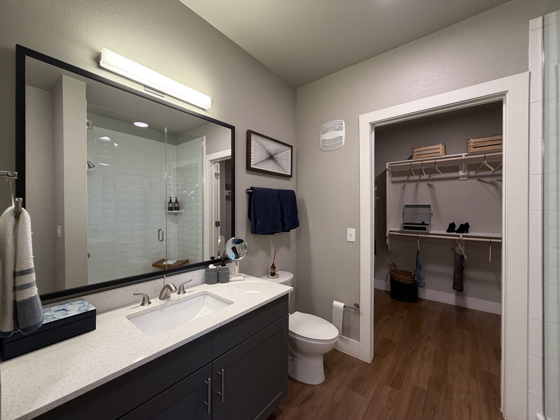 A modern bathroom features a sleek vanity with a white countertop and a rectangular sink. Above the sink, a large mirror is framed in black, reflecting the space. To the left, a glass shower with white tiled walls is visible, showcasing a minimalist design. A navy towel hangs on a rack beside the sink. The bathroom also includes a toilet and a small decorative item on the countertop. Adjacent to the bathroom is a walk-in closet with wooden shelves, hangers, and a few items, including a pair of shoes and a basket. The walls are painted in a soft gray, and the flooring is a warm wood tone.