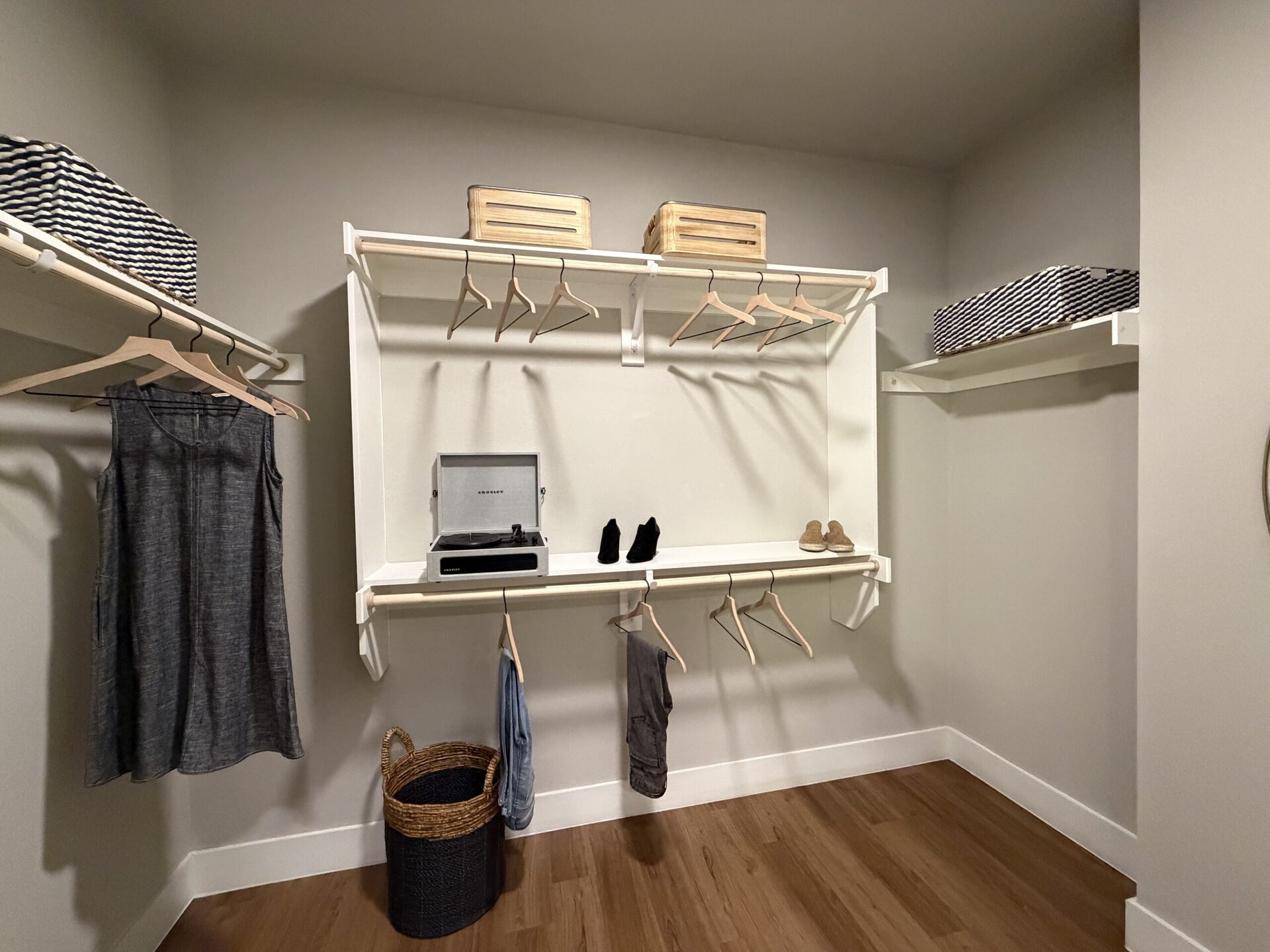 A well-organized closet features a light gray wall and wooden flooring. On the left, a gray sleeveless dress hangs from a wooden hanger. Adjacent to it, several empty hangers are positioned on a white shelf. Below, a record player sits on a shelf, accompanied by a pair of black shoes and tan sandals. Two woven baskets are placed on the upper shelves, one with a striped pattern and the other in a natural finish. A small basket is positioned on the floor, adding to the storage options.