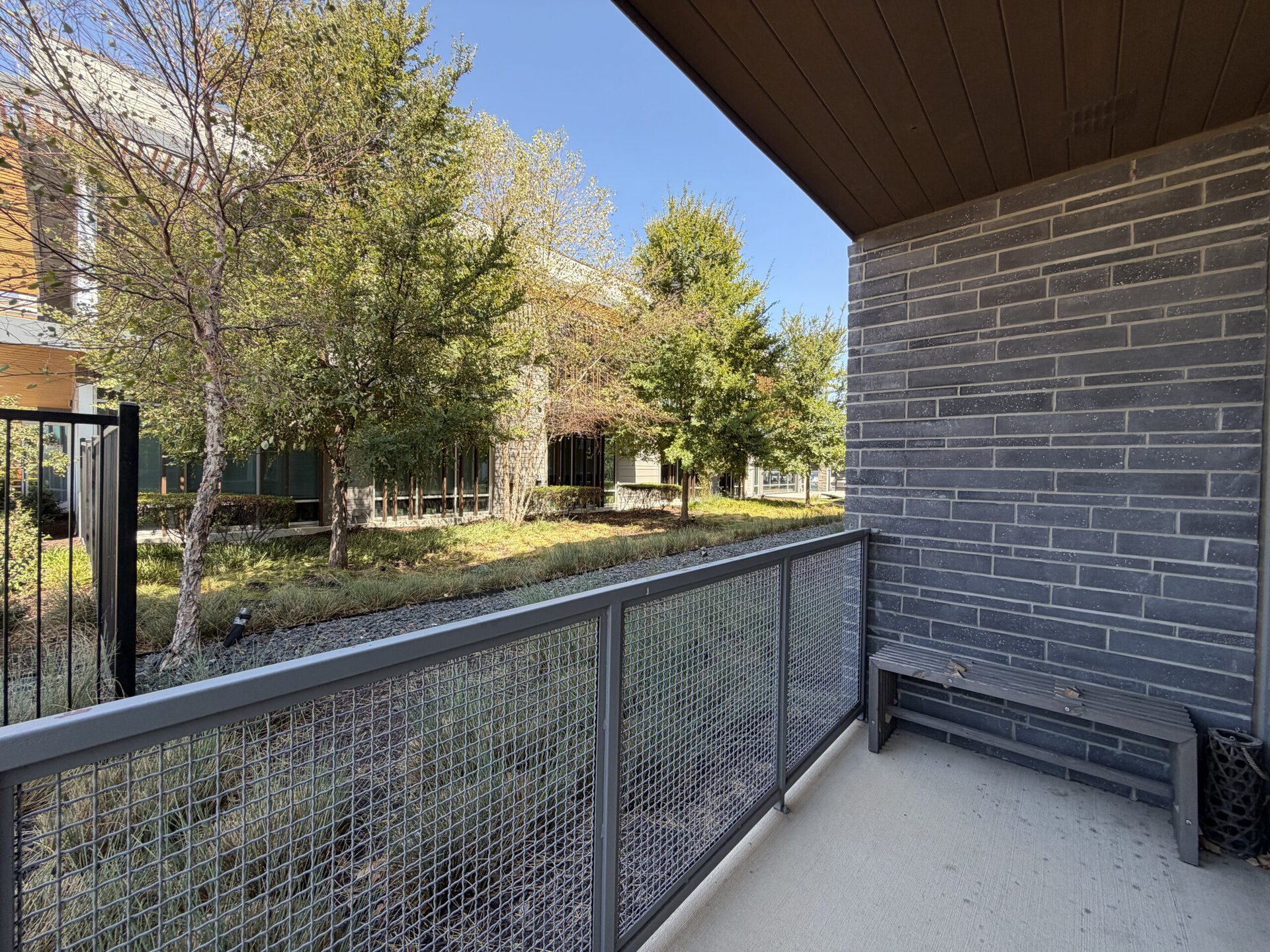 A balcony view features a gray brick wall on one side and a metal railing with a mesh design. Below, there is a wooden bench. The scene outside includes several trees and shrubs, with a pathway lined by gravel and grass. The sky is clear and blue, indicating a sunny day.
