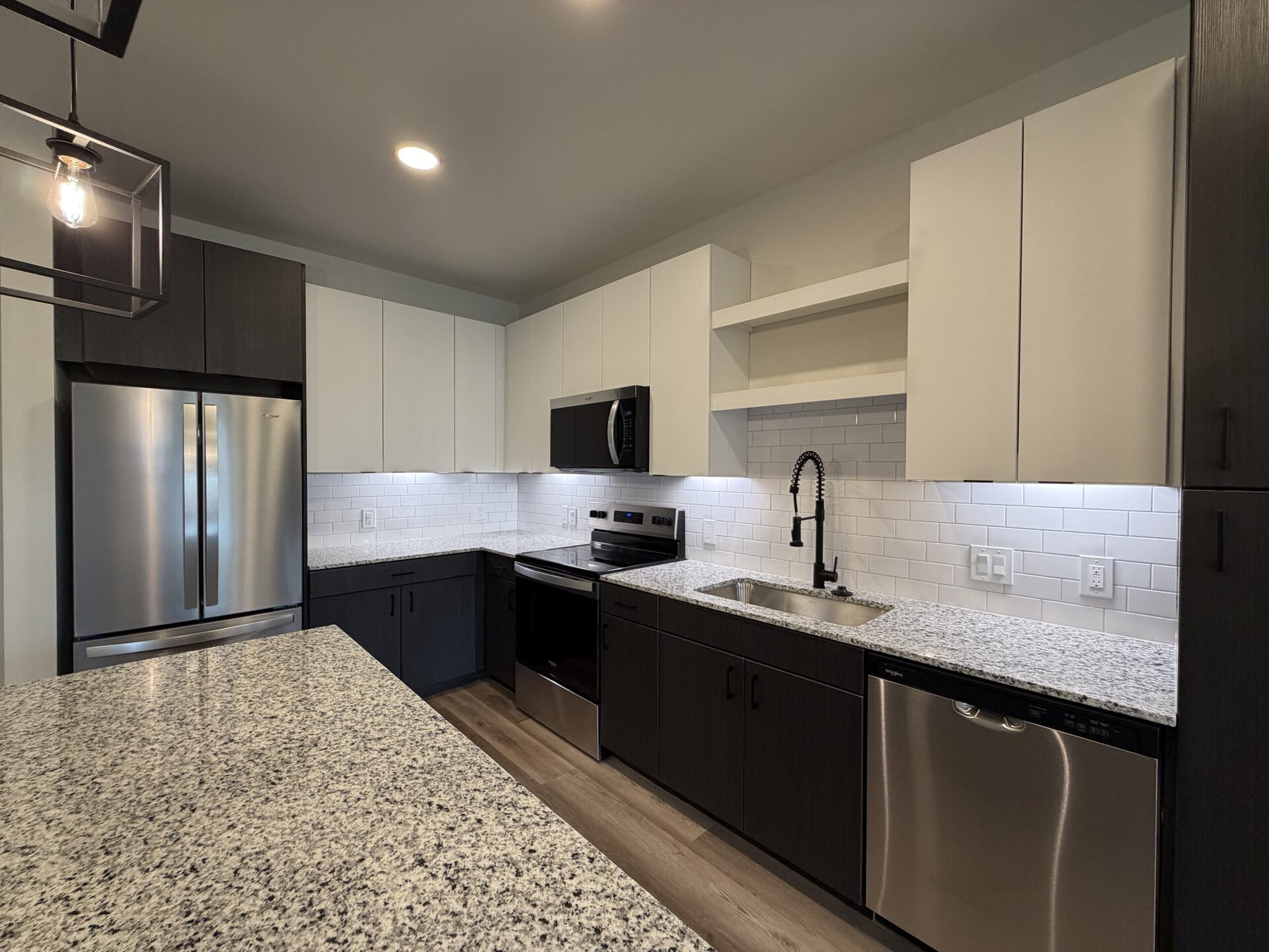 A modern kitchen featuring a combination of dark and light cabinetry. The upper cabinets are white, while the lower cabinets are a dark wood finish. A stainless steel refrigerator and microwave are integrated into the design, alongside a black stove. The countertops are made of speckled granite, and a sleek black faucet is installed above a stainless steel sink. The backsplash consists of white subway tiles, and under-cabinet lighting enhances the workspace. A pendant light hangs above the kitchen island, adding a stylish touch.