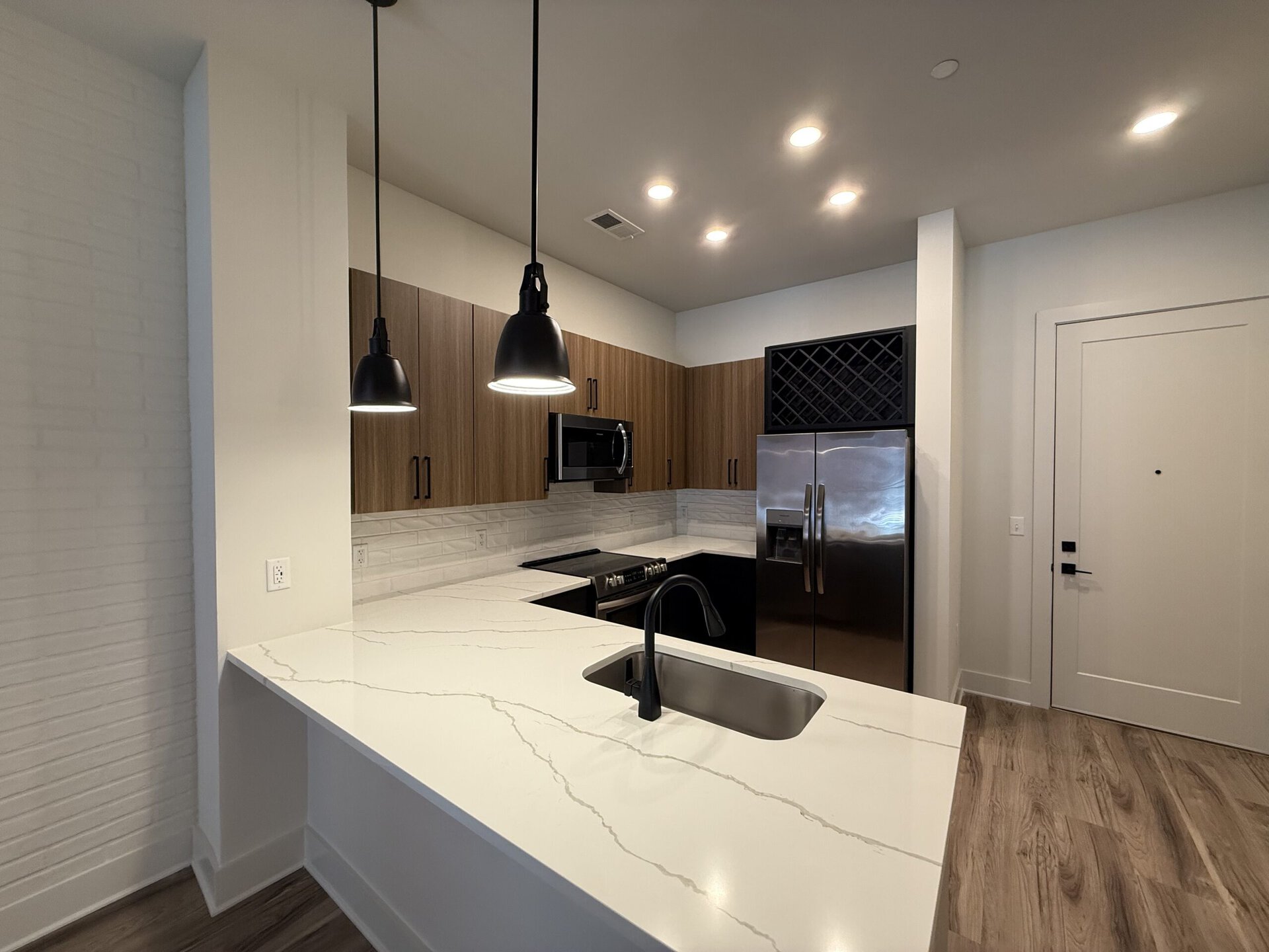 A modern kitchen featuring a large white countertop with a marble-like pattern, complemented by dark cabinetry. The kitchen includes stainless steel appliances, such as a microwave and a refrigerator with double doors. Two black pendant lights hang above the countertop, providing illumination. A sleek black faucet is installed in the sink, which is integrated into the countertop. The backsplash consists of white tiles arranged in a brick pattern, and there is a wine rack above the refrigerator. The flooring is a light wood finish, and a door is visible on the right side of the image.