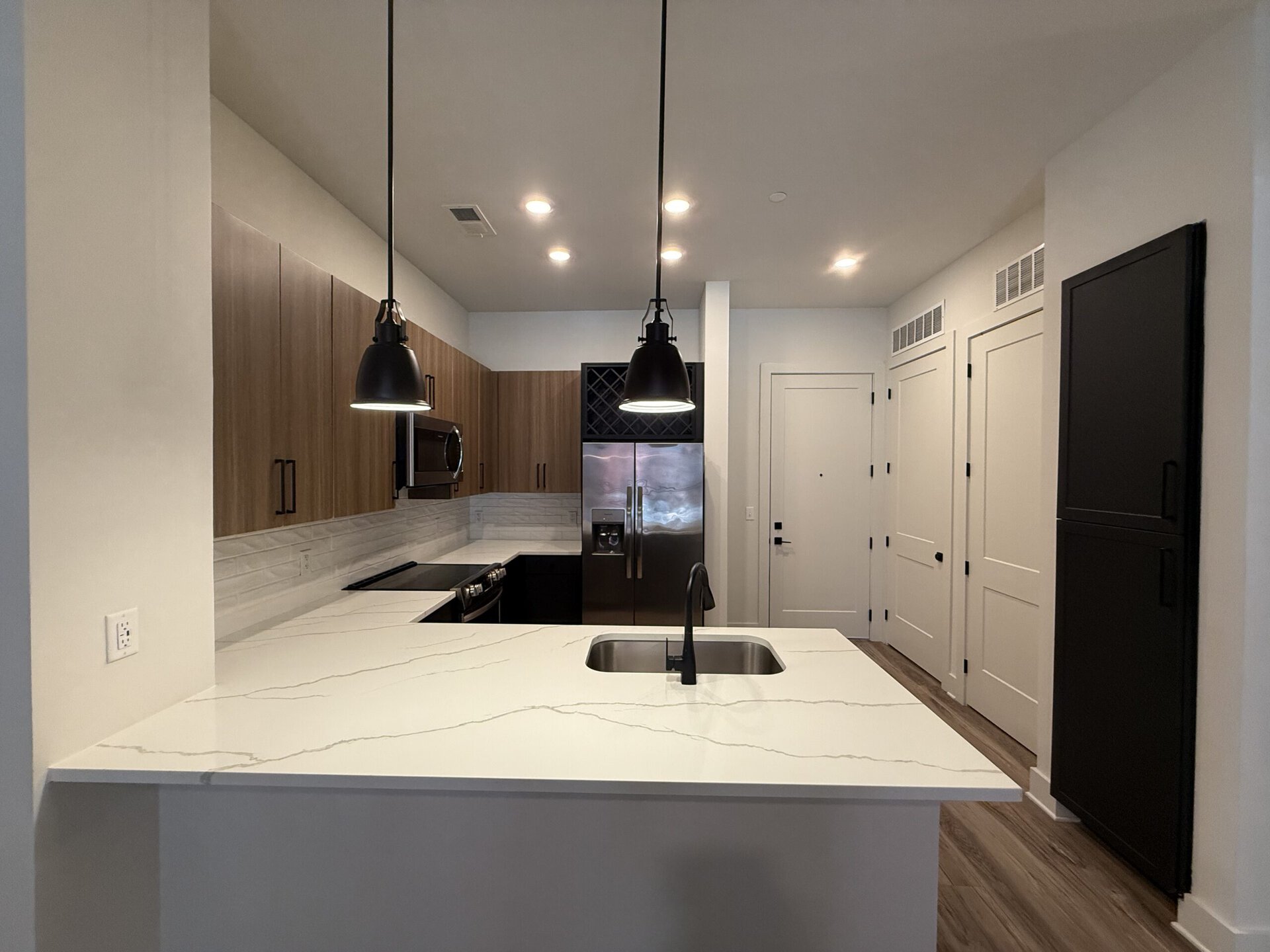 A modern kitchen featuring a large white countertop with subtle gray veining. The countertop includes a stainless steel sink and a sleek black faucet. Above, two black pendant lights hang, illuminating the space. The cabinetry is a mix of light wood and dark finishes, with a stainless steel refrigerator and built-in microwave visible. The backsplash consists of white tiles, and the flooring is a warm wood tone. A doorway leads to a hallway with additional doors, suggesting storage or utility spaces.