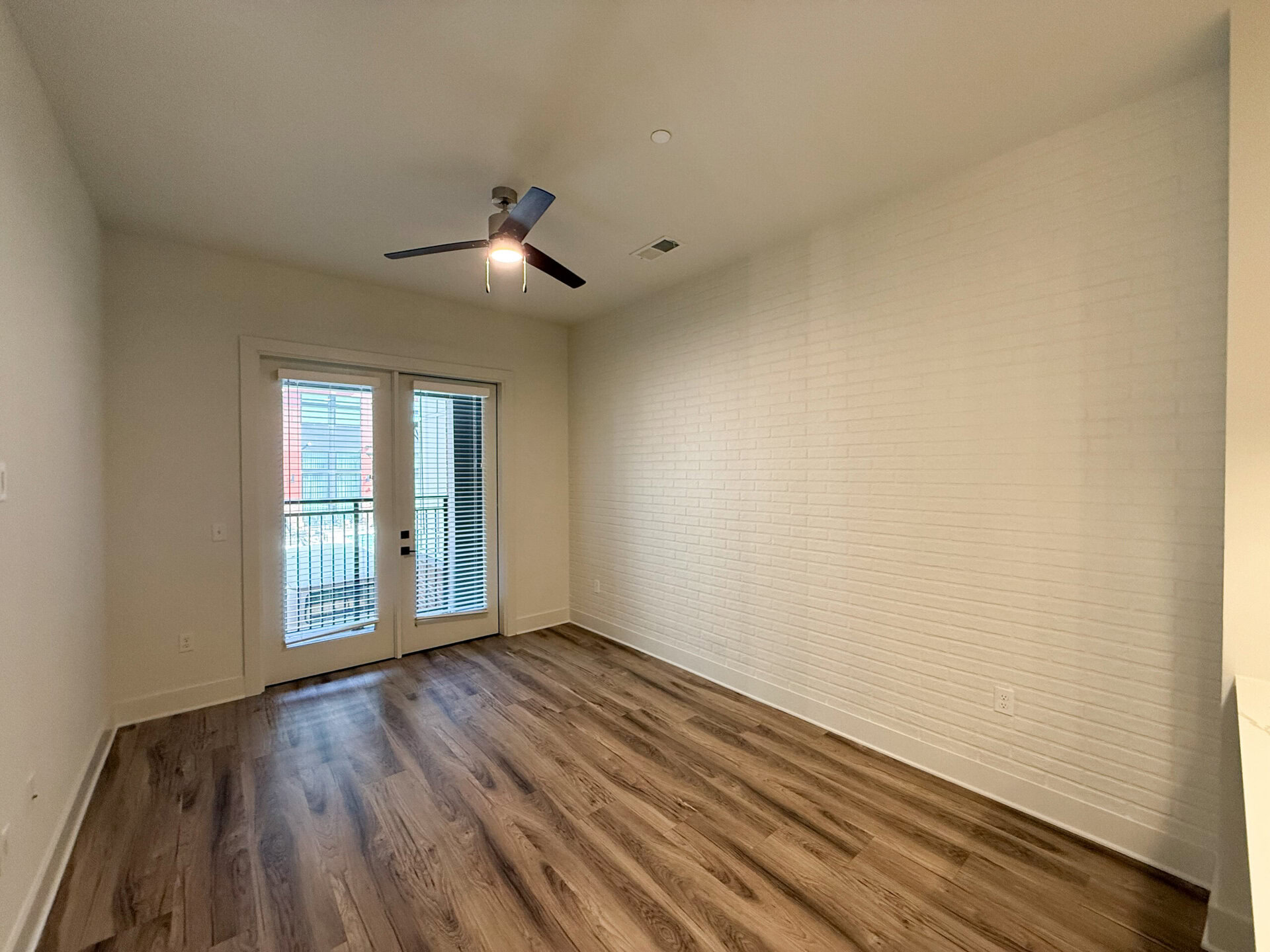 A spacious room features light-colored walls with a textured white brick accent wall. The flooring is a warm-toned wood laminate. A ceiling fan with a light fixture is mounted above. Large double doors with blinds lead to a balcony, allowing natural light to fill the space. The overall ambiance is bright and modern, with minimal furnishings.