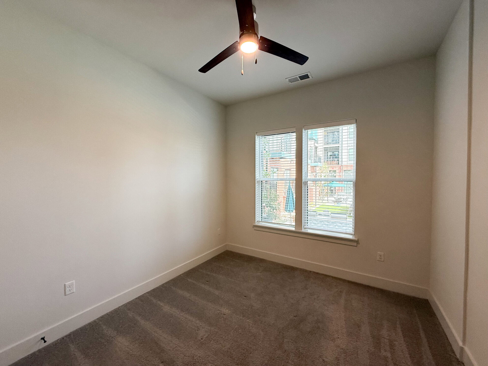 A small, empty room features light-colored walls and a plush carpeted floor. A ceiling fan with dark blades hangs above, providing a modern touch. Two large windows with white blinds allow natural light to enter, revealing a view of an outdoor area with greenery and furniture. The room has a clean and minimalistic appearance, ideal for various uses.