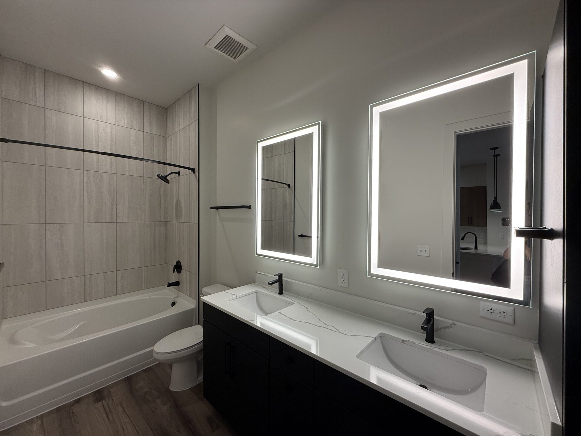 A modern bathroom features a sleek design with light gray tiled walls and a bathtub on the left. The vanity area includes a white countertop with two sinks, accented by a striking black faucet. Above the sinks, two illuminated mirrors provide ample lighting. A black towel bar is mounted on the wall, and the overall aesthetic is clean and contemporary. The space is well-lit with recessed lighting.