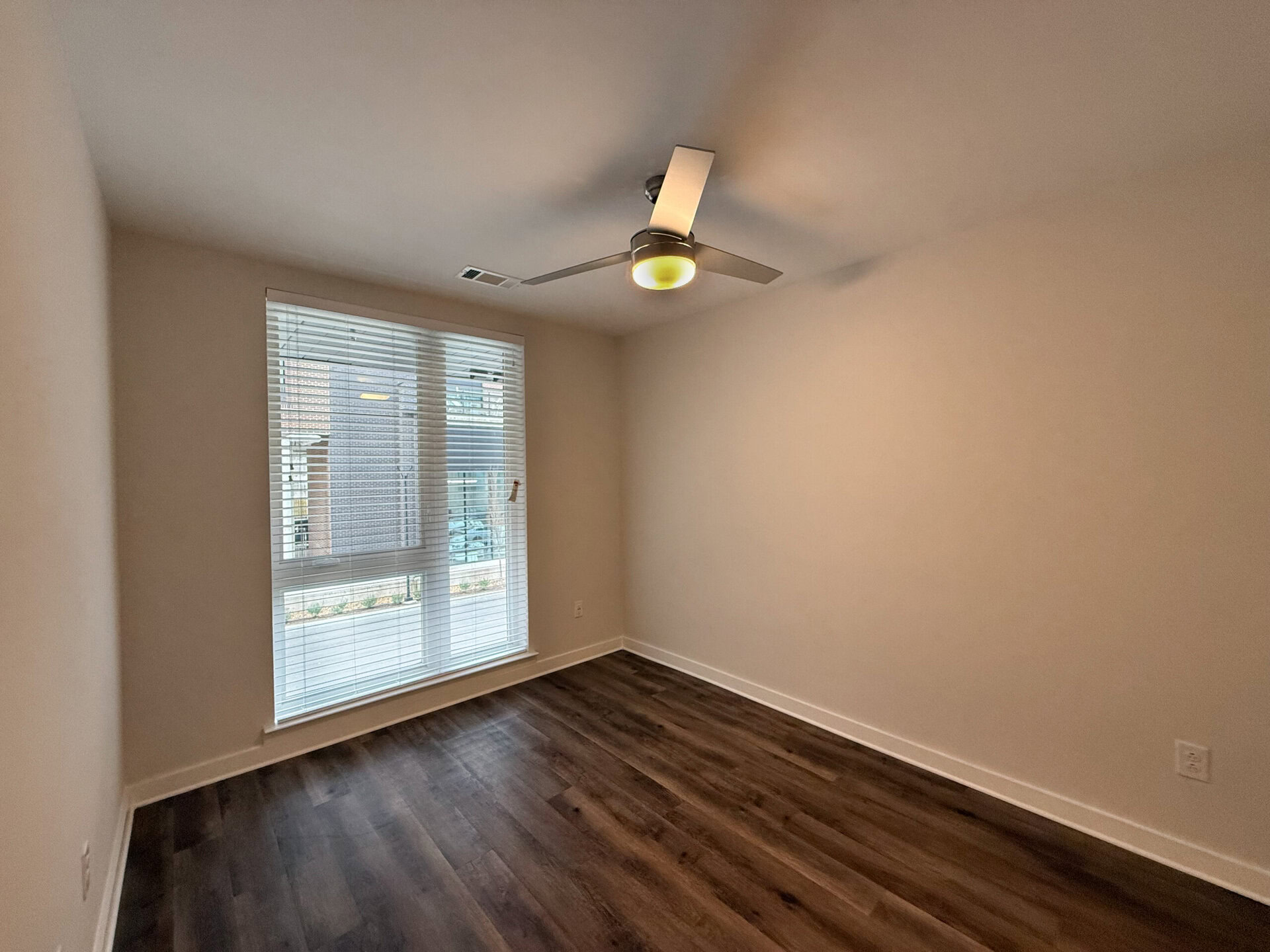 A small, empty room features light-colored walls and a dark wood floor. A ceiling fan with a light fixture is mounted in the center. On one side, a large window with horizontal blinds allows natural light to enter, revealing a view of the outside. The overall atmosphere is bright and minimalistic, with ample space for furniture.