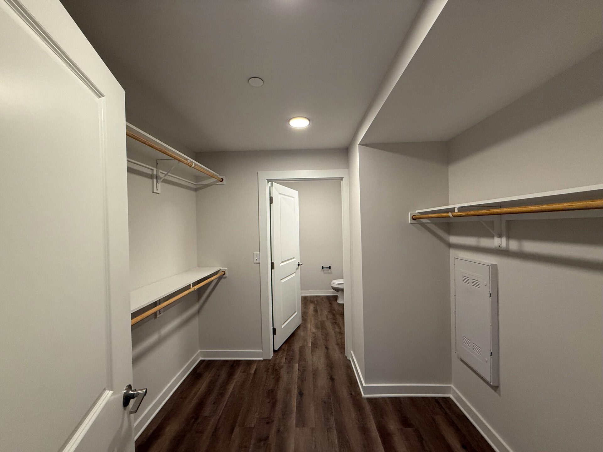 A spacious walk-in closet features light gray walls and a wood-like laminate floor. On the left side, there are two wooden hanging rods mounted on brackets, providing ample space for clothing. A shelf runs beneath the rods. The closet opens to a doorway leading to a bathroom, which is partially visible, showing a toilet. The ceiling is equipped with recessed lighting, enhancing the bright and airy feel of the space.