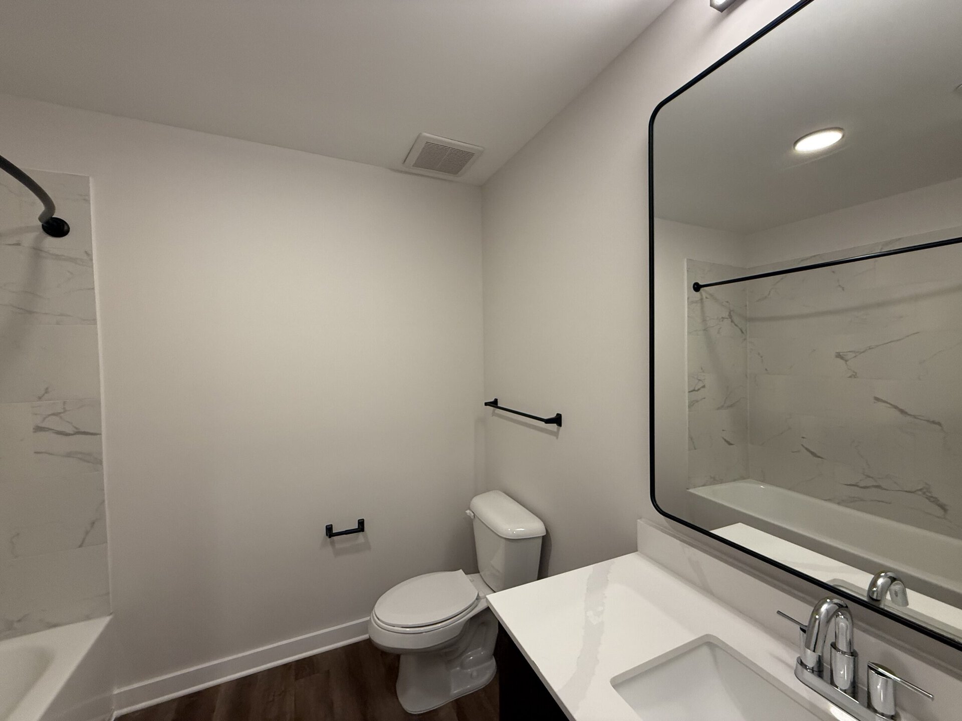 A modern bathroom features a clean design with light-colored walls. On the left, there is a shower area with a marble-patterned wall and a black shower rod. A white bathtub is visible in the background. To the right, a sleek white countertop with a chrome faucet sits beneath a large, rounded black-framed mirror. A white toilet is positioned nearby, with two black towel bars mounted on the walls. The overall aesthetic is minimalist and contemporary.