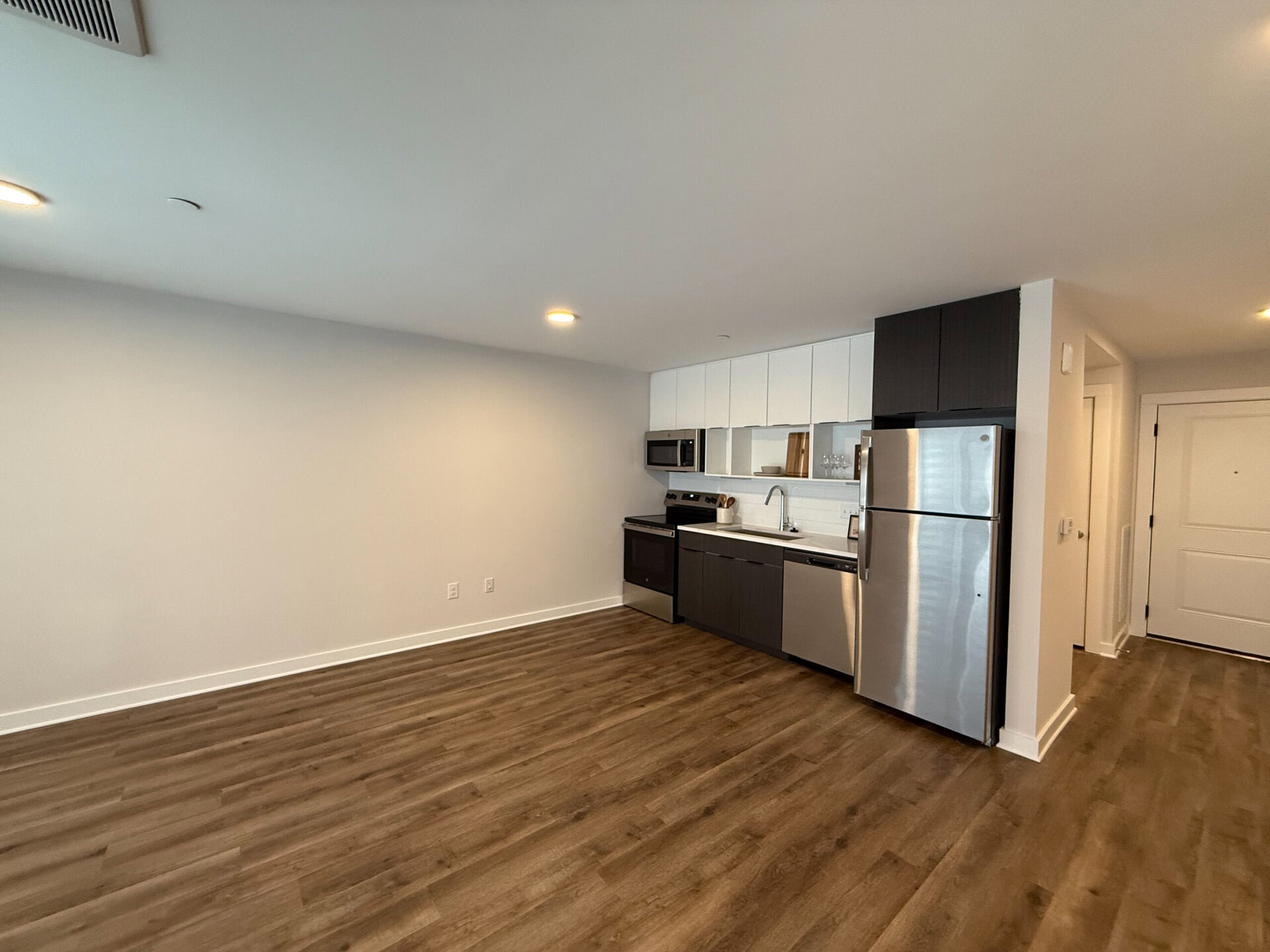 A modern kitchen area features sleek cabinetry with a mix of white and dark finishes. Stainless steel appliances include a refrigerator, microwave, and oven. A stainless steel sink is positioned under a window, with a countertop that extends along the wall. The flooring is a warm, wood-like laminate, and the walls are painted in a light neutral color. A door is visible on the right, leading to another area, while the space is well-lit with recessed lighting.