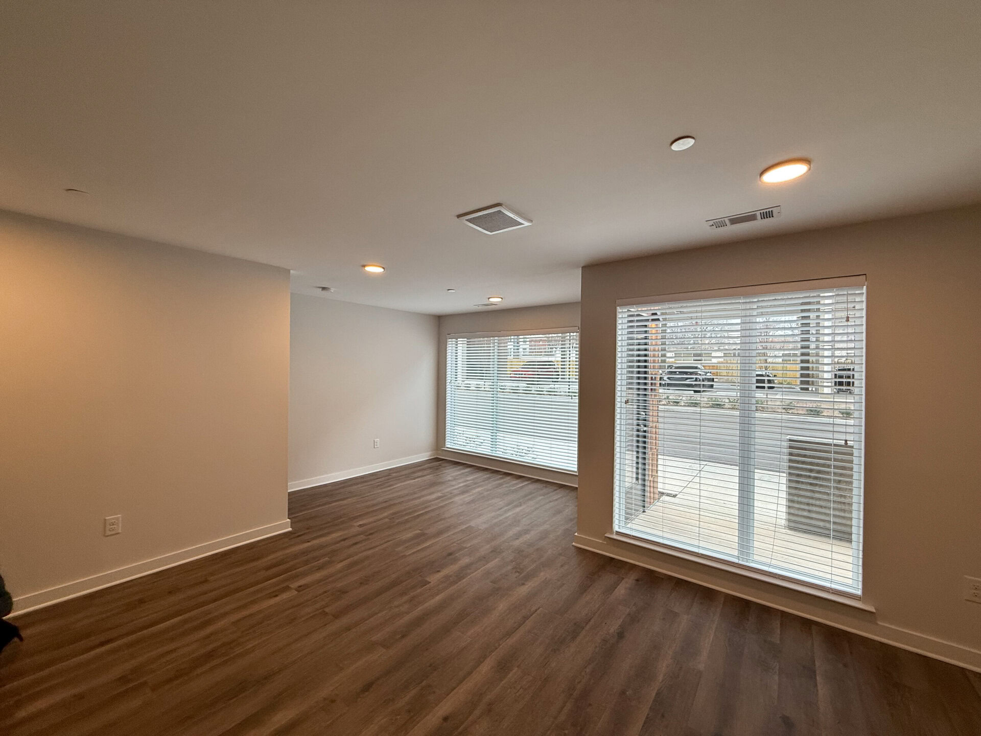 A spacious, well-lit living area features light-colored walls and a warm wooden floor. Large windows with white blinds allow natural light to fill the room, and there is a door leading outside. The ceiling is equipped with recessed lighting, enhancing the modern feel of the space. The overall atmosphere is open and inviting, suitable for various furniture arrangements.