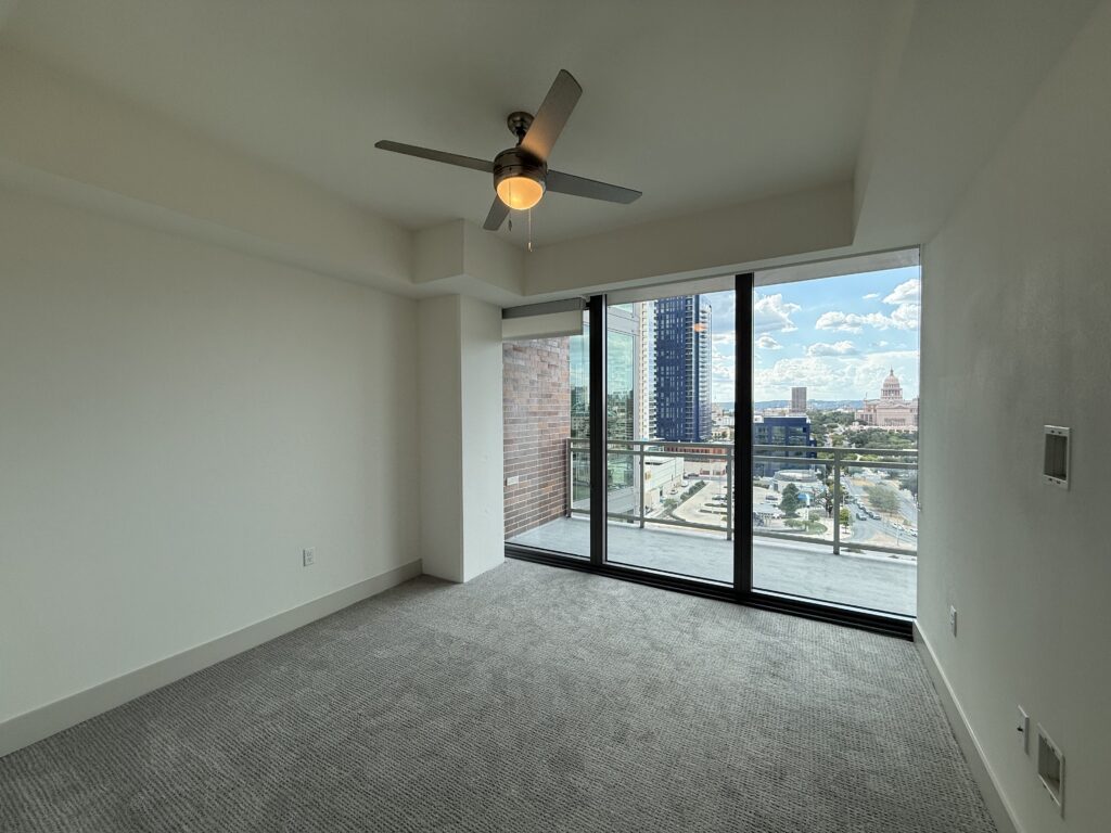 A spacious room features a light-colored wall and plush carpet. A ceiling fan with a light fixture hangs from the ceiling. Large glass doors lead to a balcony, offering a view of a cityscape with modern buildings and a distant landmark. The room is well-lit with natural light streaming in through the glass doors.