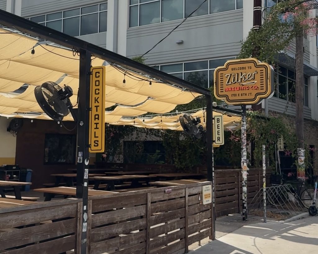 An outdoor seating area of a brewery features wooden picnic-style tables under a canopy of beige fabric. Two large fans are mounted to provide airflow. Prominent signs indicate "COCKTAILS" and "BEER," while a larger sign reads "WELCOME TO ZILKER BREWING CO." The background includes a building with large windows and greenery, adding to the inviting atmosphere.
