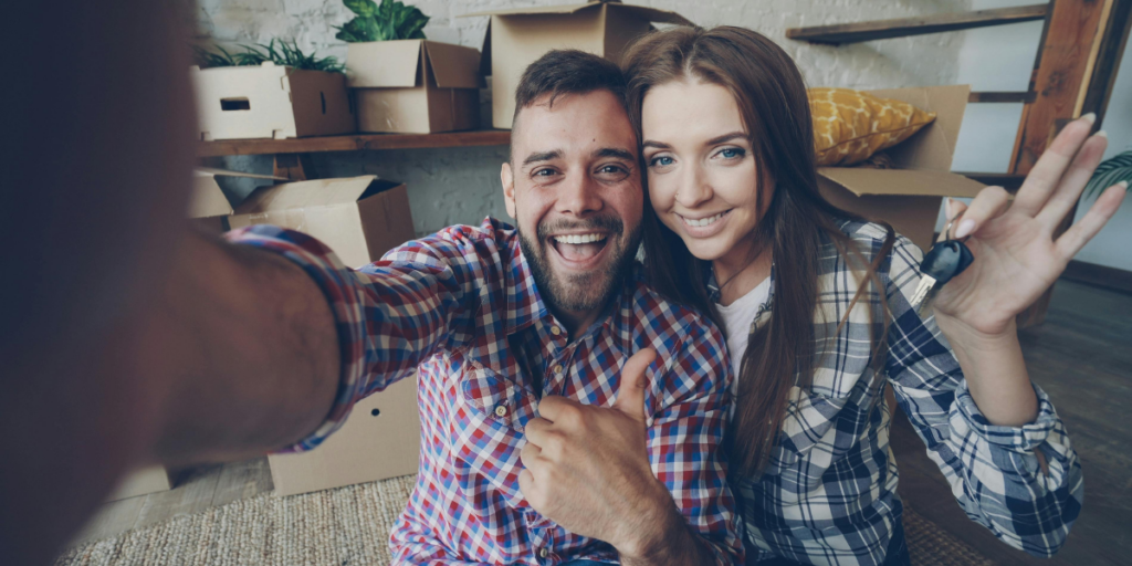 A smiling first-time renter couple takes a selfie in a room filled with cardboard boxes, suggesting a recent move. The man, wearing a plaid shirt, gives a thumbs-up, while the woman, in a checkered shirt, smiles and holds a set of keys. The background features a few plants and a yellow cushion on a shelf, adding a cozy touch to the space.