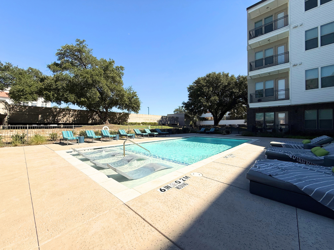 A clear blue sky overlooks a swimming pool surrounded by a spacious deck. The pool features a shallow entry area and is bordered by several lounge chairs with turquoise cushions. To one side, there are large, comfortable loungers with striped fabric and bright green accents. In the background, a multi-story building with balconies is visible, along with a large tree providing shade. The area is well-maintained, with landscaping elements enhancing the outdoor space.