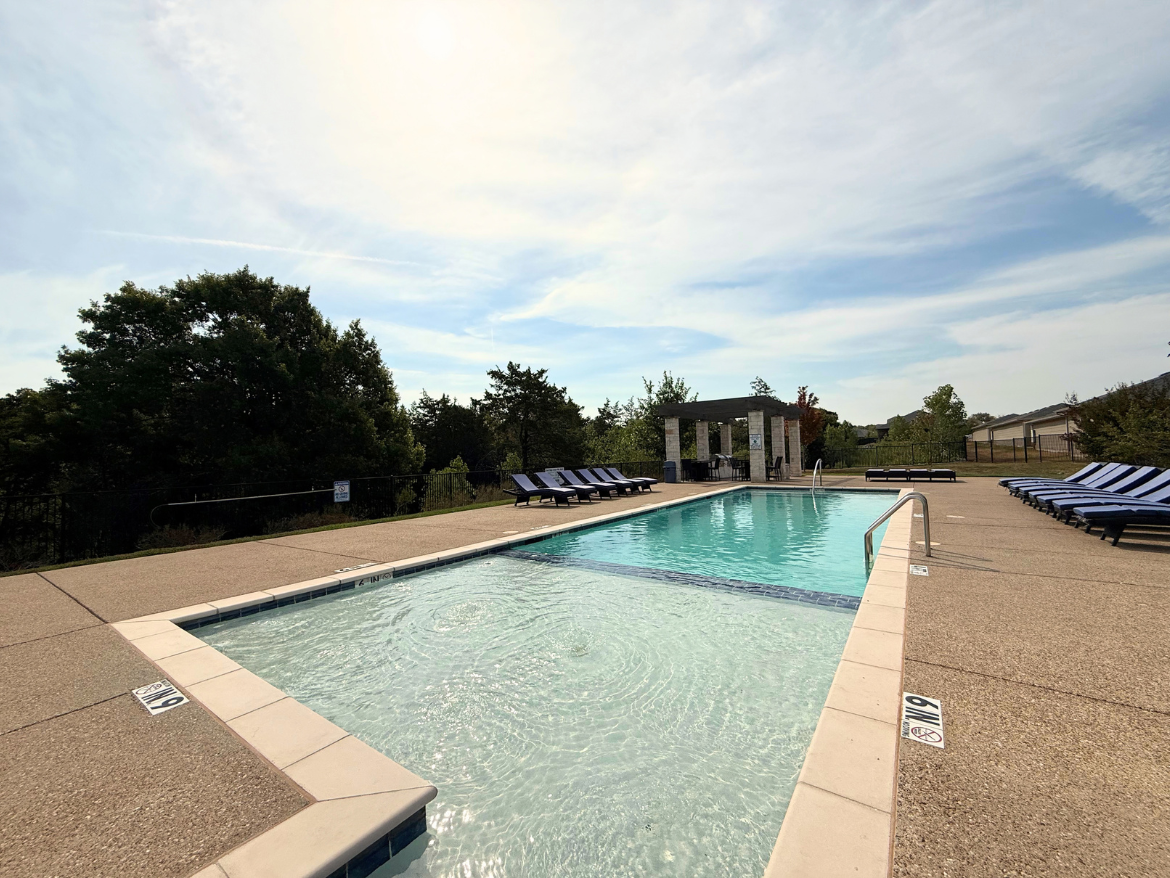 A rectangular swimming pool is visible, featuring clear blue water and a light-colored concrete deck. On one side, there are several lounge chairs with blue and white striped cushions arranged neatly. In the background, there is a small shaded pavilion with stone columns and greenery surrounding the area. The sky is partly cloudy, creating a bright and inviting atmosphere.