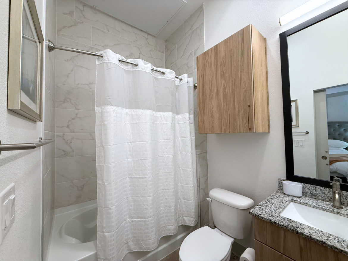 A modern bathroom features a white shower curtain hanging from a silver rod, partially concealing a bathtub with a curved edge. The walls are tiled with light-colored marble, and a wooden cabinet is mounted above a granite countertop. A rectangular mirror with a black frame reflects part of the room, and a white sink is positioned next to a sleek faucet. A toilet is situated beside the sink, completing the compact layout.