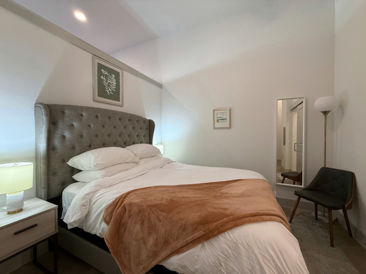 A cozy bedroom features a large, tufted gray headboard with white bedding and a soft, light brown throw blanket draped over the bed. On either side of the bed, there are small nightstands with lamps. A framed botanical print hangs above the headboard, and a full-length mirror is positioned next to a stylish chair with a dark green upholstery. The room has a warm, inviting atmosphere with soft lighting and neutral colors.