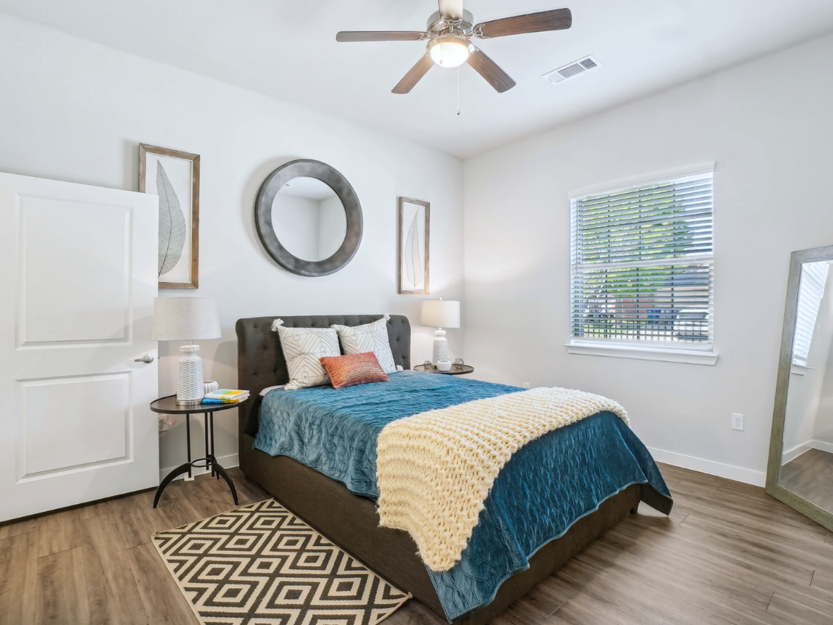 A cozy bedroom features a plush bed with a teal blanket and a chunky cream-colored throw. The headboard is upholstered in dark fabric, and decorative pillows add a pop of color. A round mirror hangs above the bed, flanked by two framed leaf prints. A small side table with a lamp sits next to the bed, accompanied by a few books. Natural light streams through a window with blinds, and a large mirror stands against one wall, reflecting the room's warm tones and wooden flooring.
