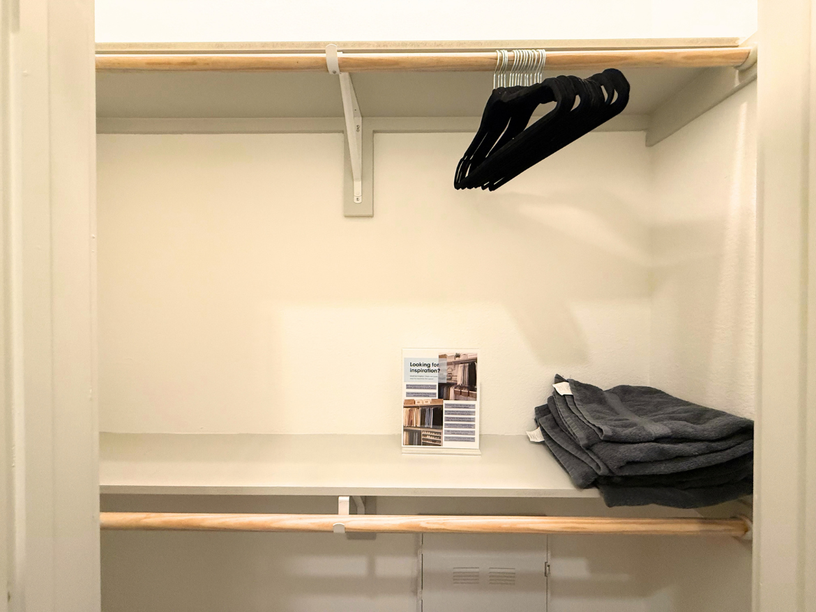 An empty closet features a wooden shelf and a hanging rod with several black hangers. On the shelf, there is a stack of neatly folded gray towels. A small brochure titled "Looking for inspiration?" is displayed upright on the shelf. The walls are painted a light color, contributing to a clean and organized appearance.